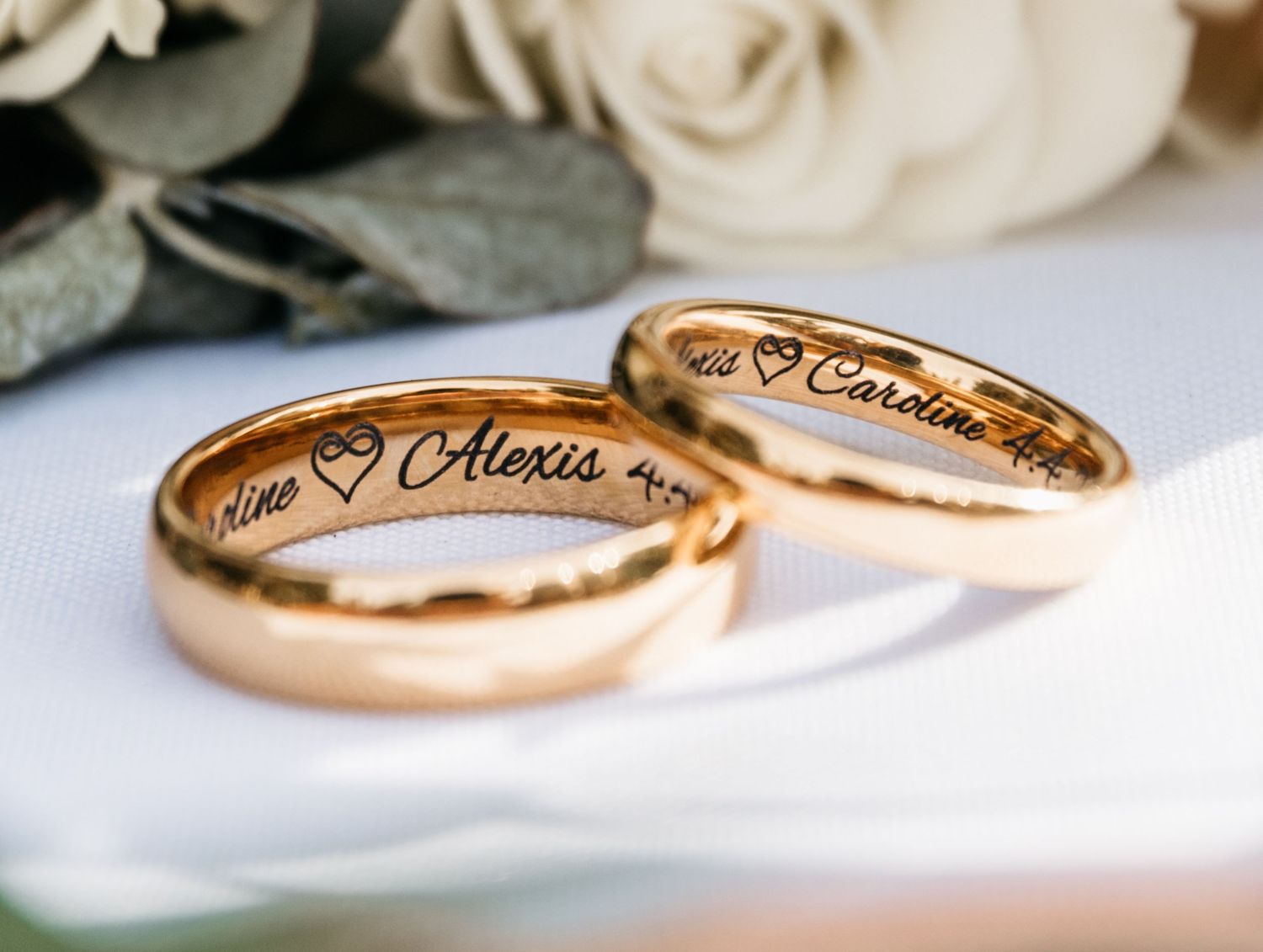 Two gold wedding bands with engraved inscriptions displayed on white rose petals.