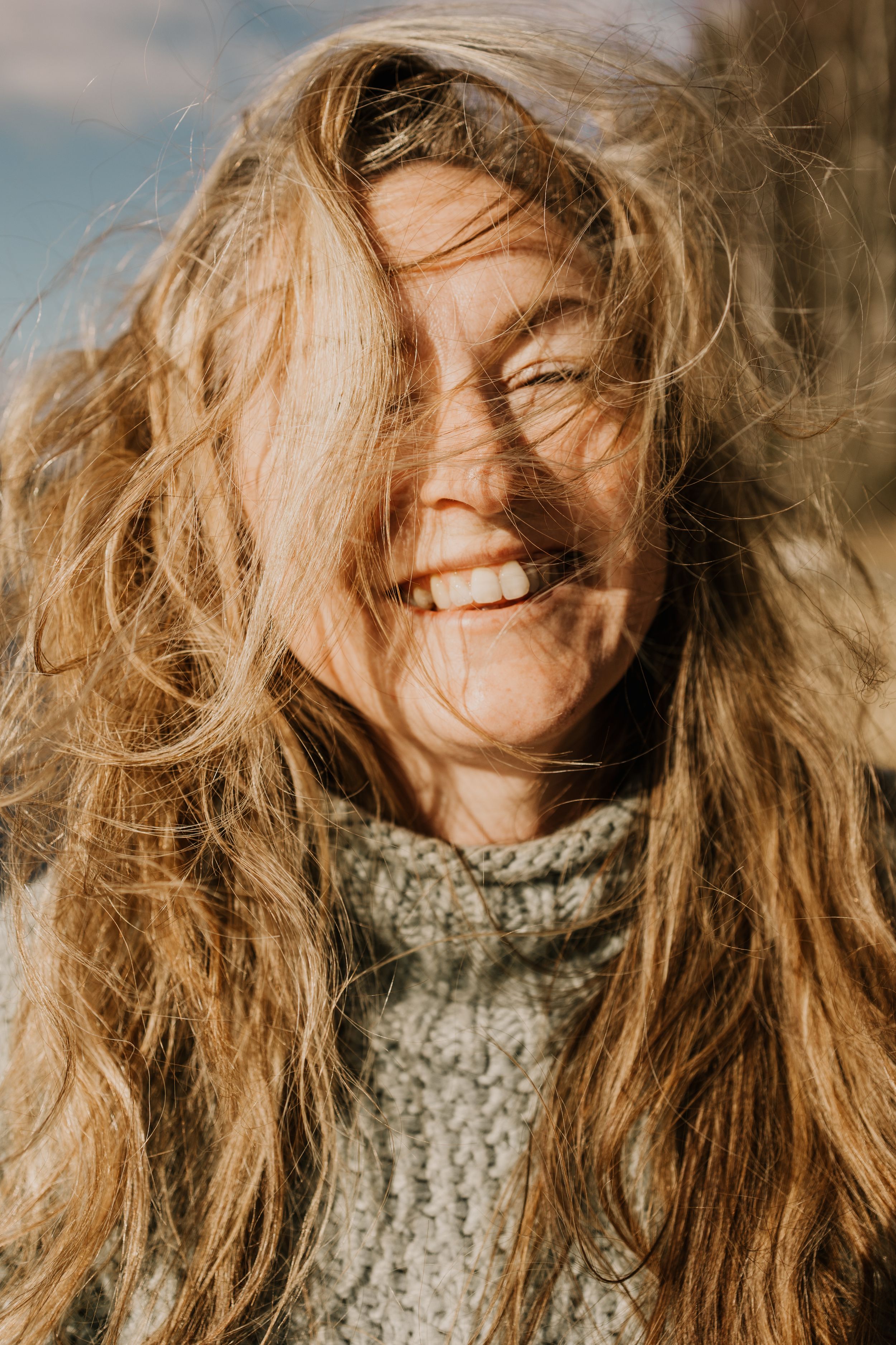 Long wavy blonde hair flows in sunlight as someone in a knit sweater smiles warmly during golden hour.