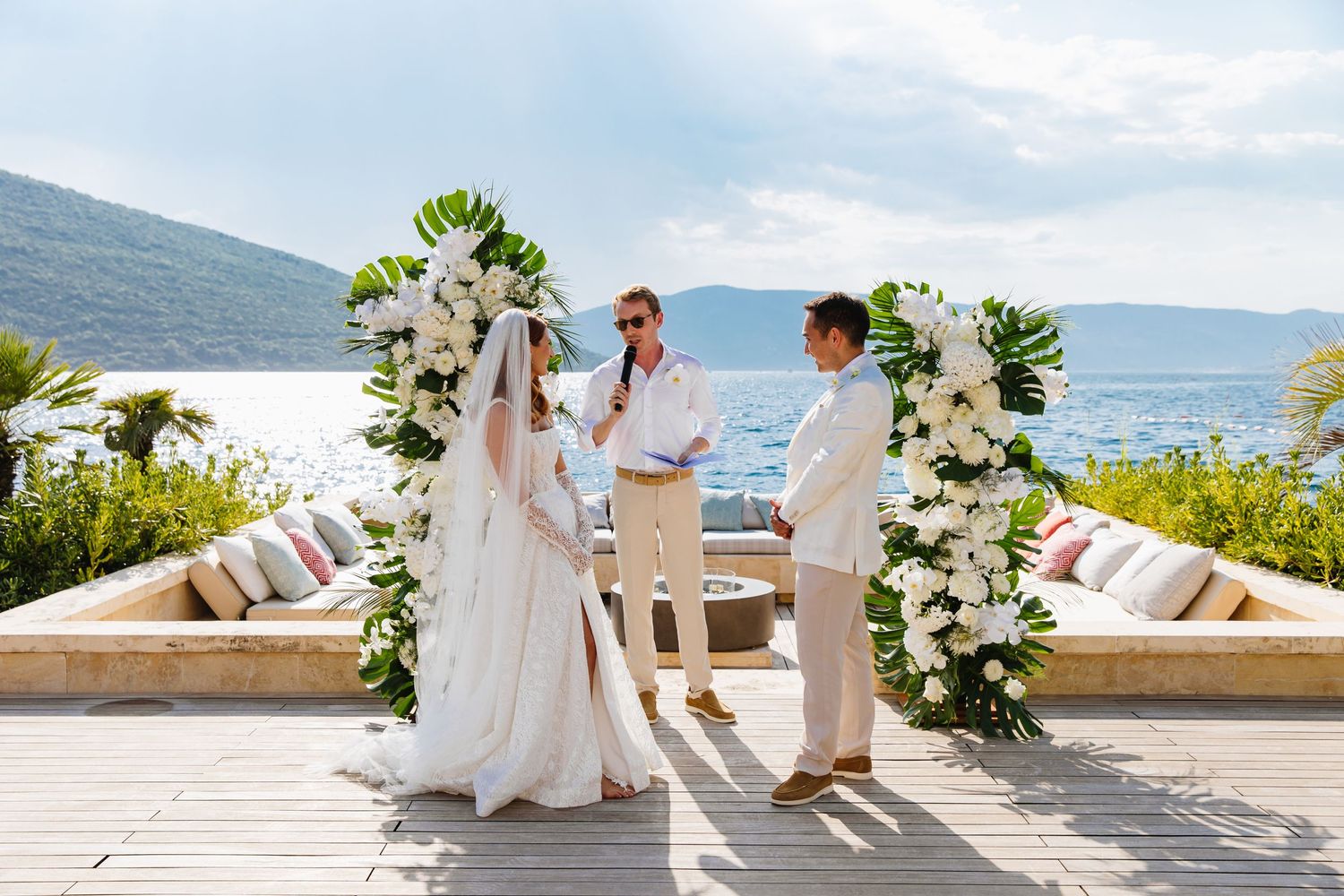 Seaside wedding ceremony setup with white floral arch and mountain views.