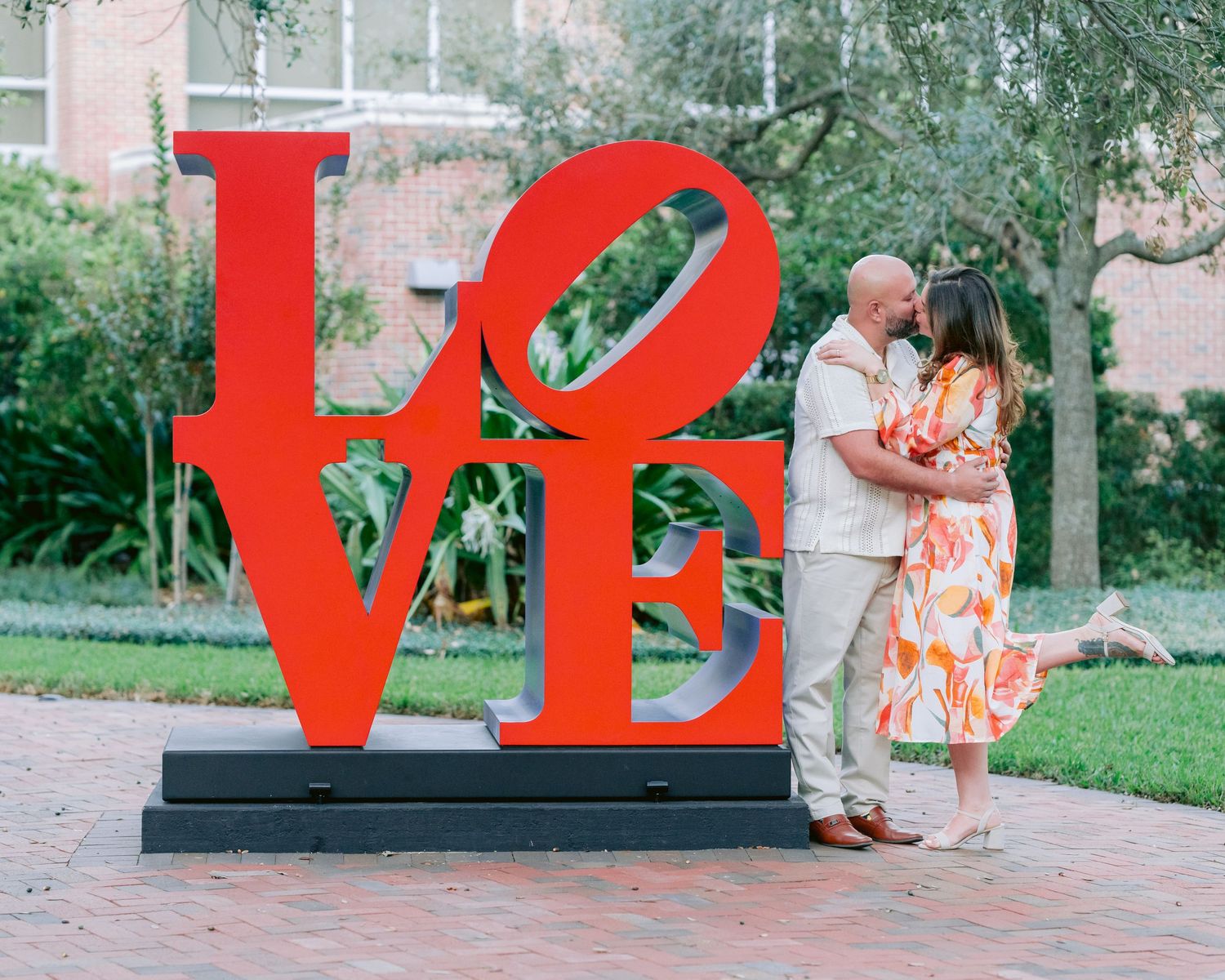 A couple embraces in front of the iconic red LOVE sculpture in a park garden setting.