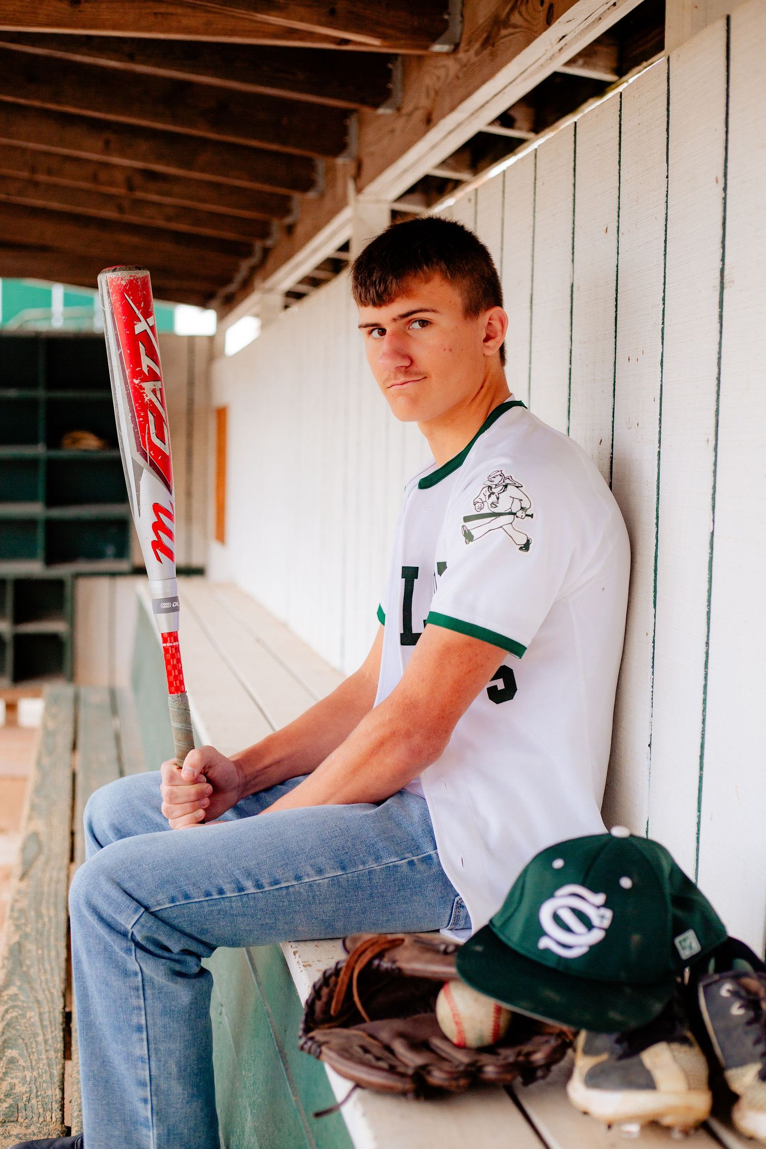 Boy senior Baseball player in white uniform sits in dugout with equipment including bat, glove and green team cap for senior photos with NSP