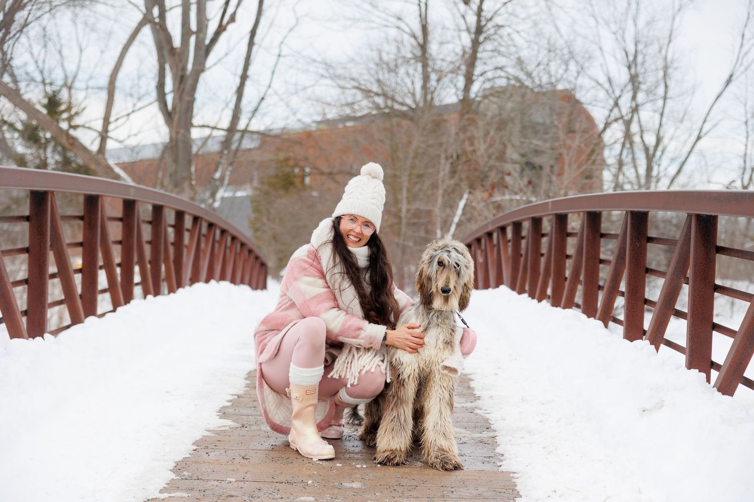 A person in a pink winter coat and white beanie poses with their dog on a snowy wooden bridge during winter.