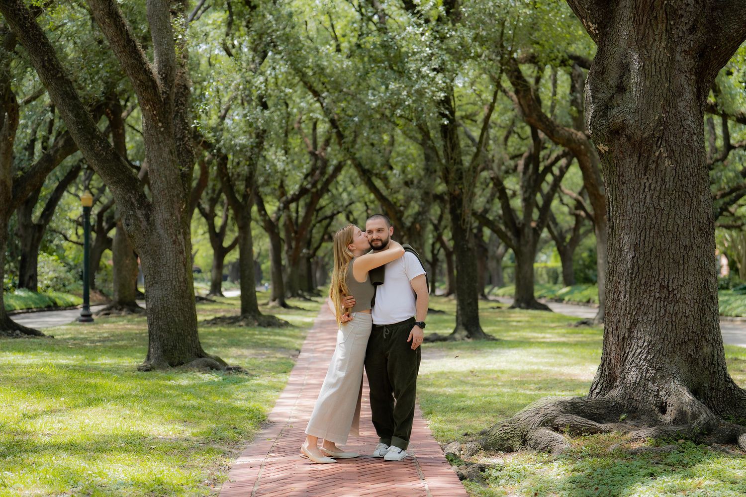 A couple walks together on a brick path under a canopy of majestic oak trees in a peaceful garden setting.