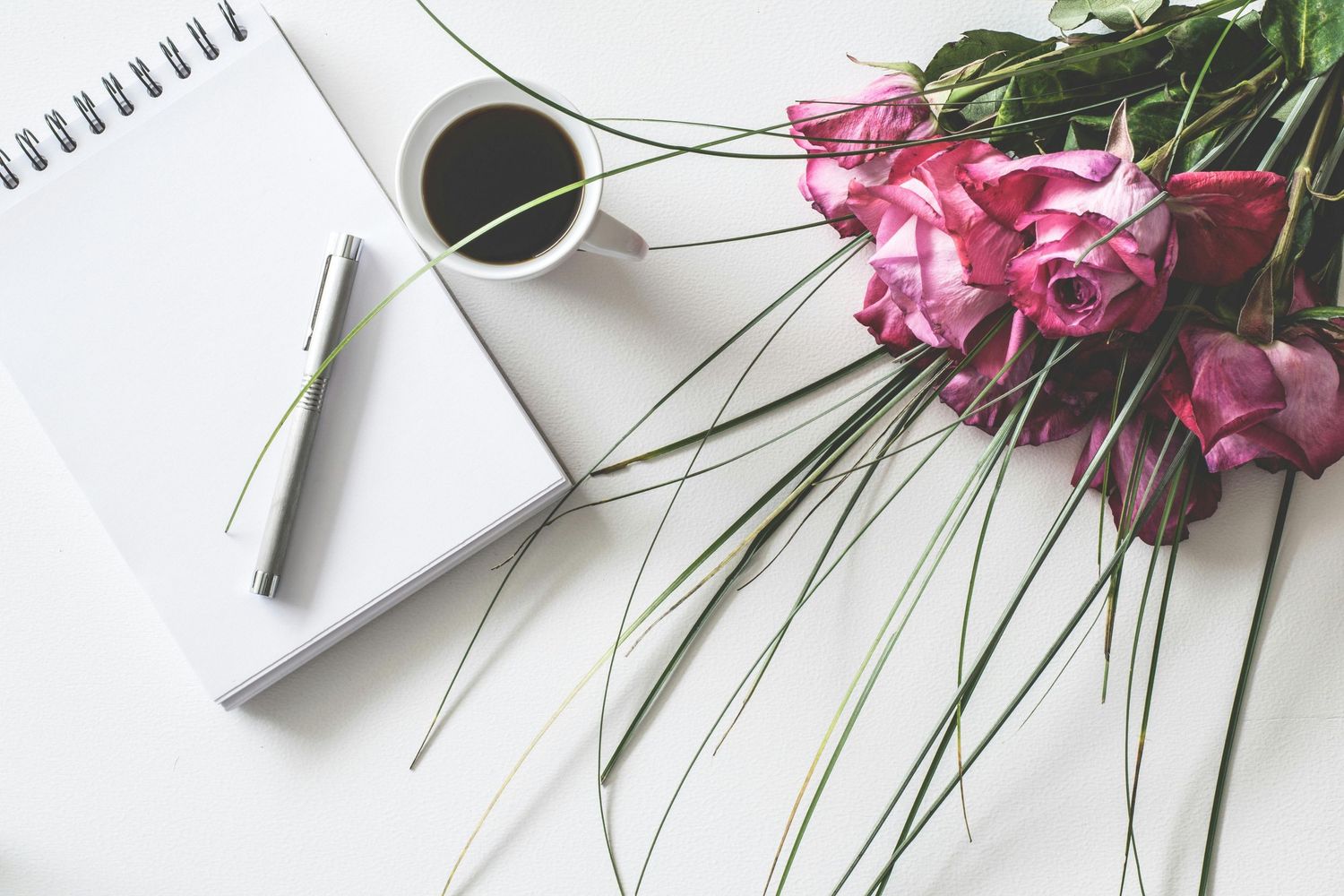 Blank notebook with coffee cup and pink flowers on white surface with modern minimalist styling.