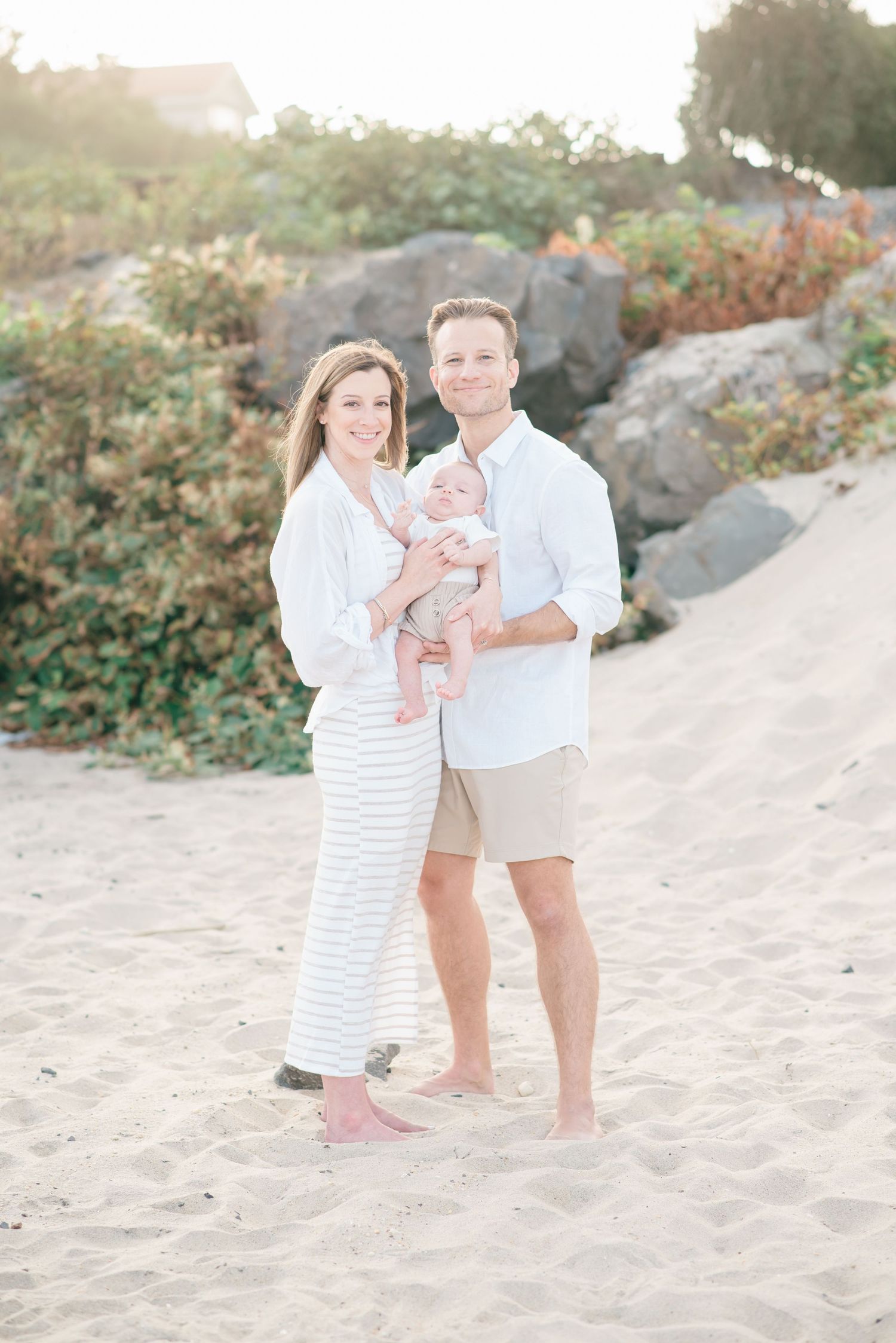 A couple that searched photographer near me is holding their one month old son in front of greenery on deal beach.