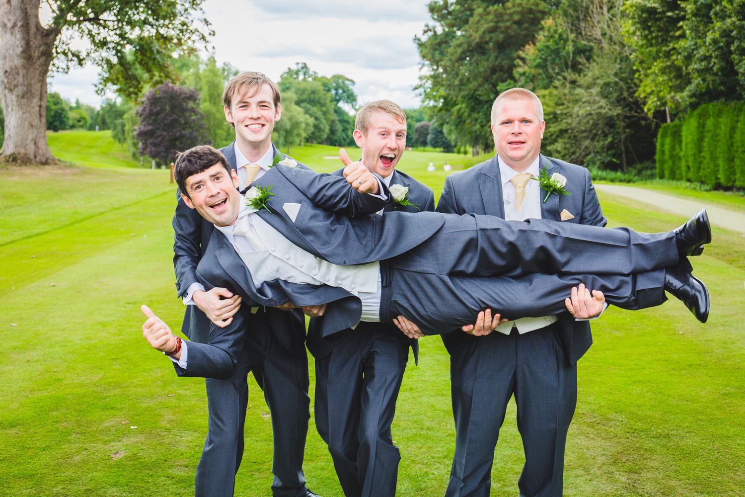 Groomsmen in dark suits playfully carrying someone across a green golf course.