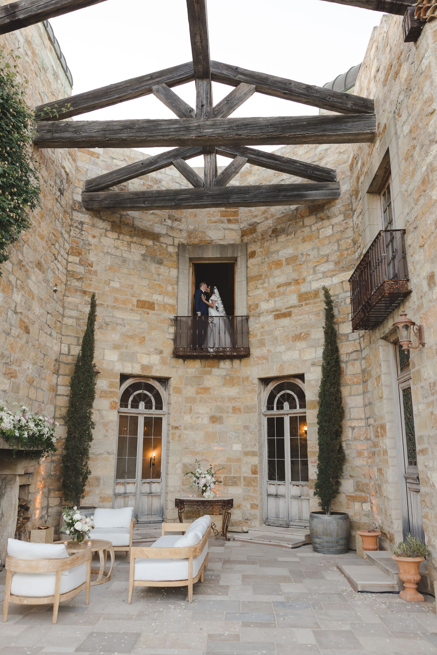 Rustic stone courtyard with wooden beams, elegant arched windows, cypress trees and outdoor lounge furniture at a luxury venue.