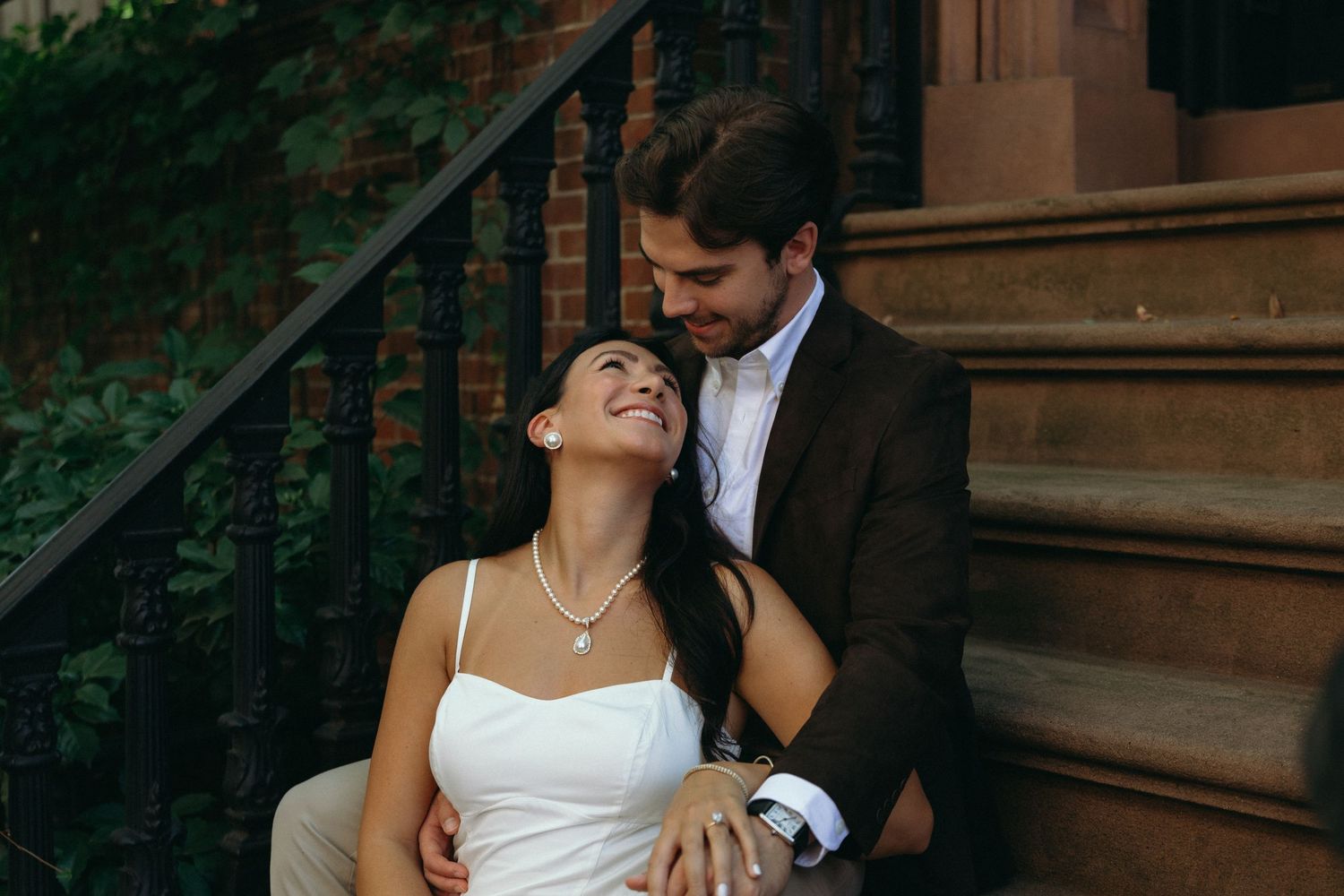 A romantic couple in wedding attire share an intimate moment on brownstone steps with greenery in the background.