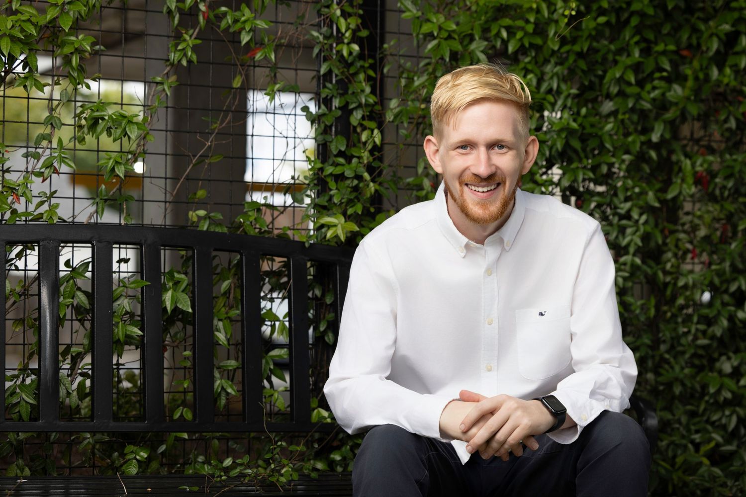 Chef in white uniform smiles while posing against green vine-covered wall background.