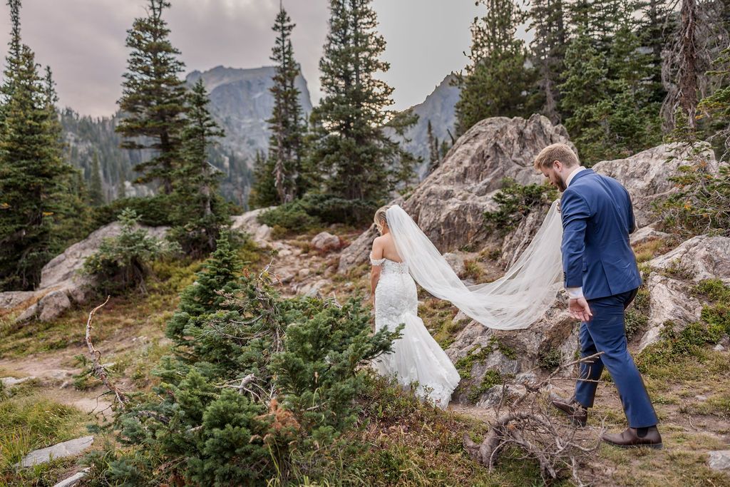A couple in wedding attire poses on a scenic mountain trail surrounded by evergreen trees and dramatic peaks.