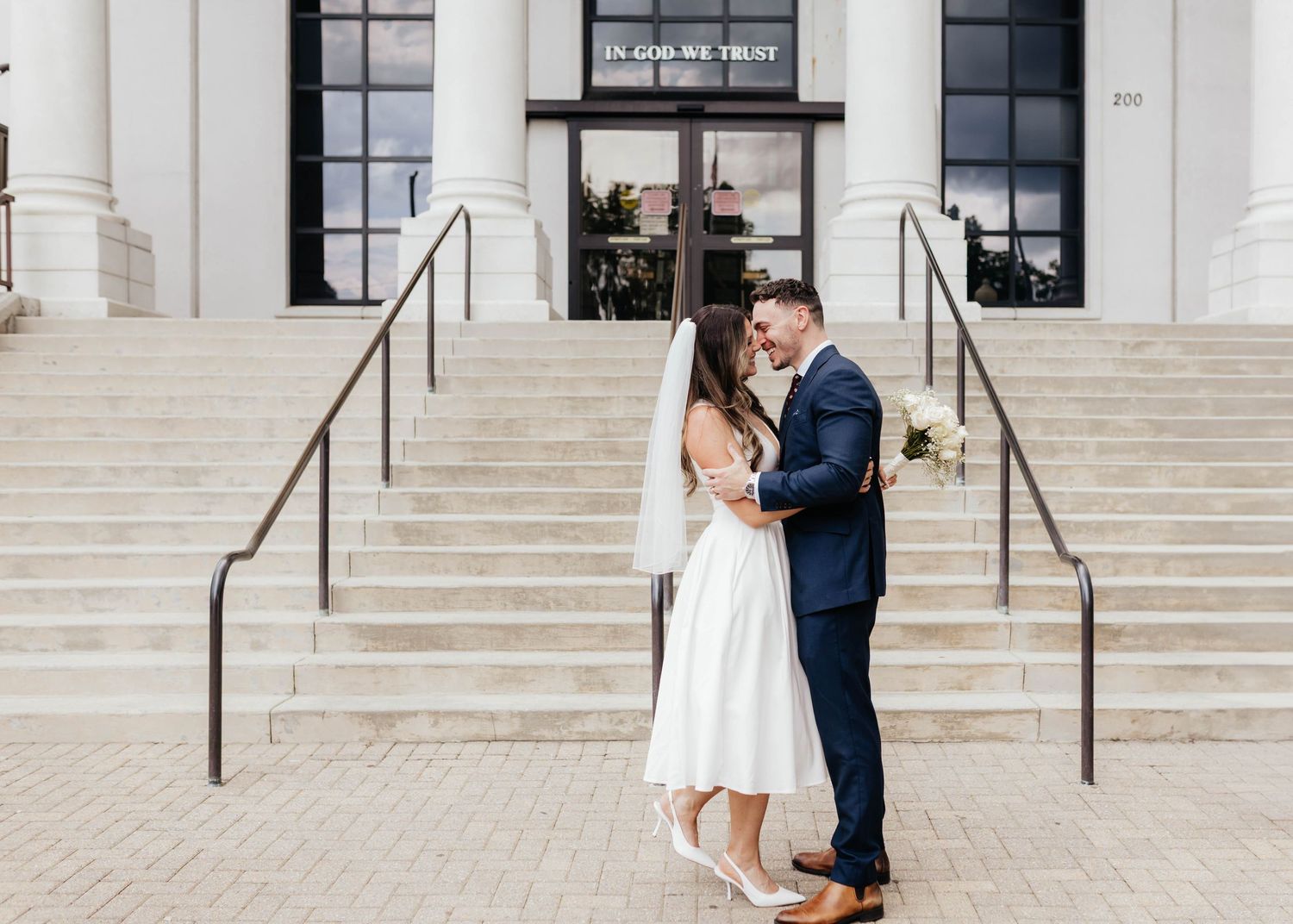 A couple in wedding attire embrace on the steps of a white building with columns and handrails.