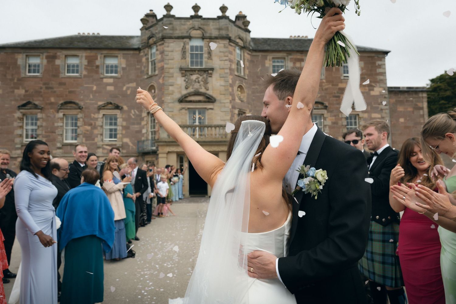 Couple walking through a garden path in wedding attire on a rainy day with umbrellas.