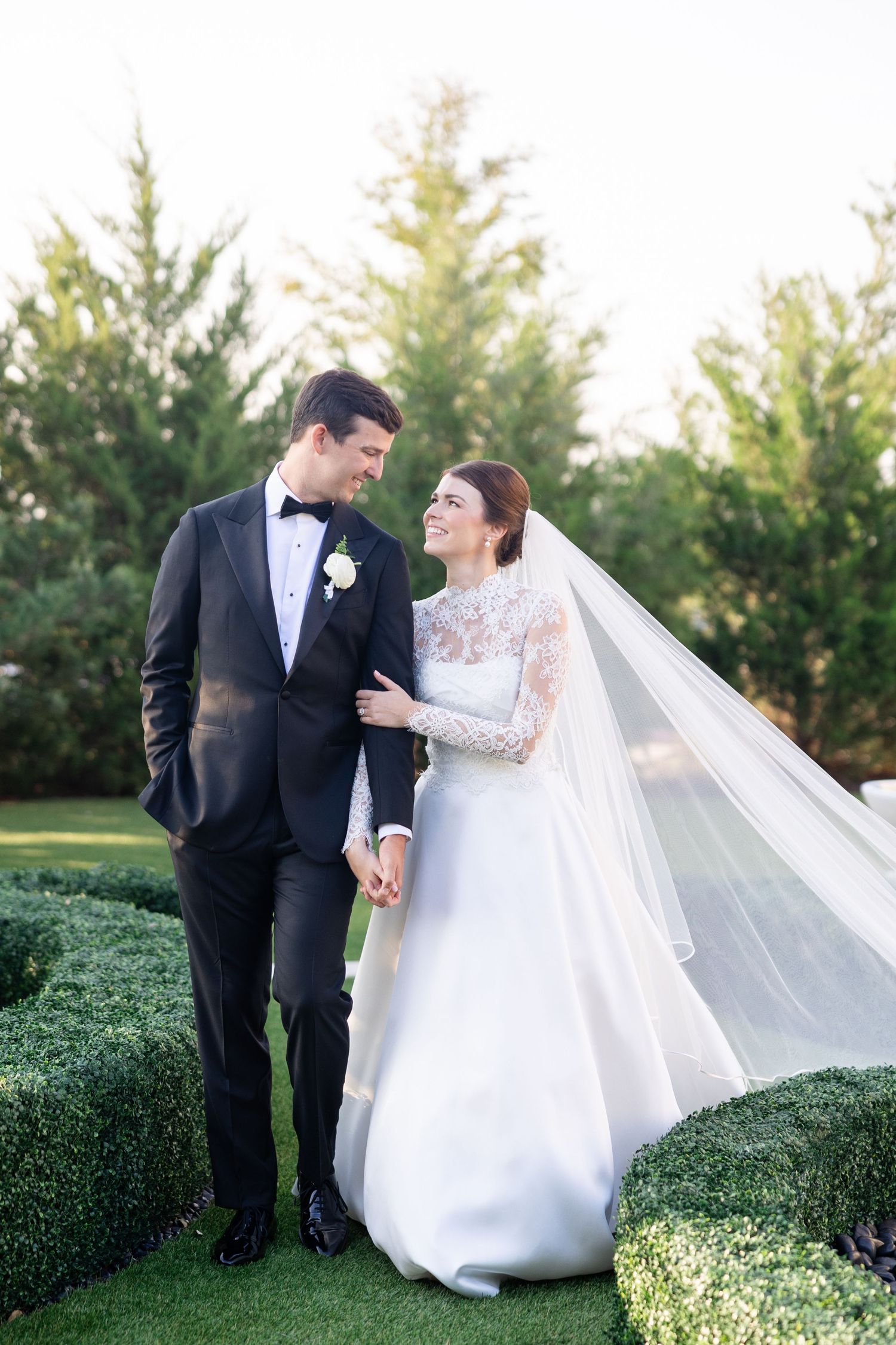 A bride and groom share a romantic moment in a lush garden, she in a lace wedding gown with veil, he in a black tuxedo.