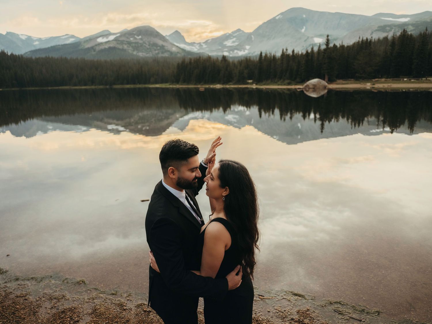A romantic couple takes wedding photos at sunset by a serene lakeside with snow-capped mountains in the background.