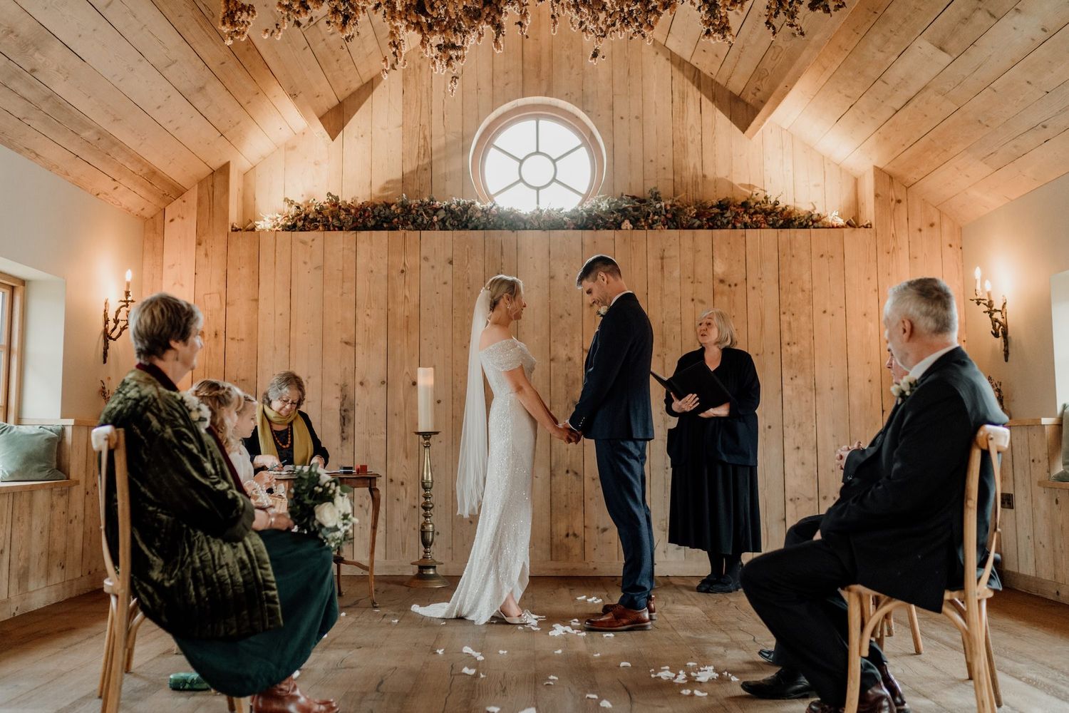 Wedding party gathers in a rustic wooden barn venue with white decor and warm lighting for multiple photographs.