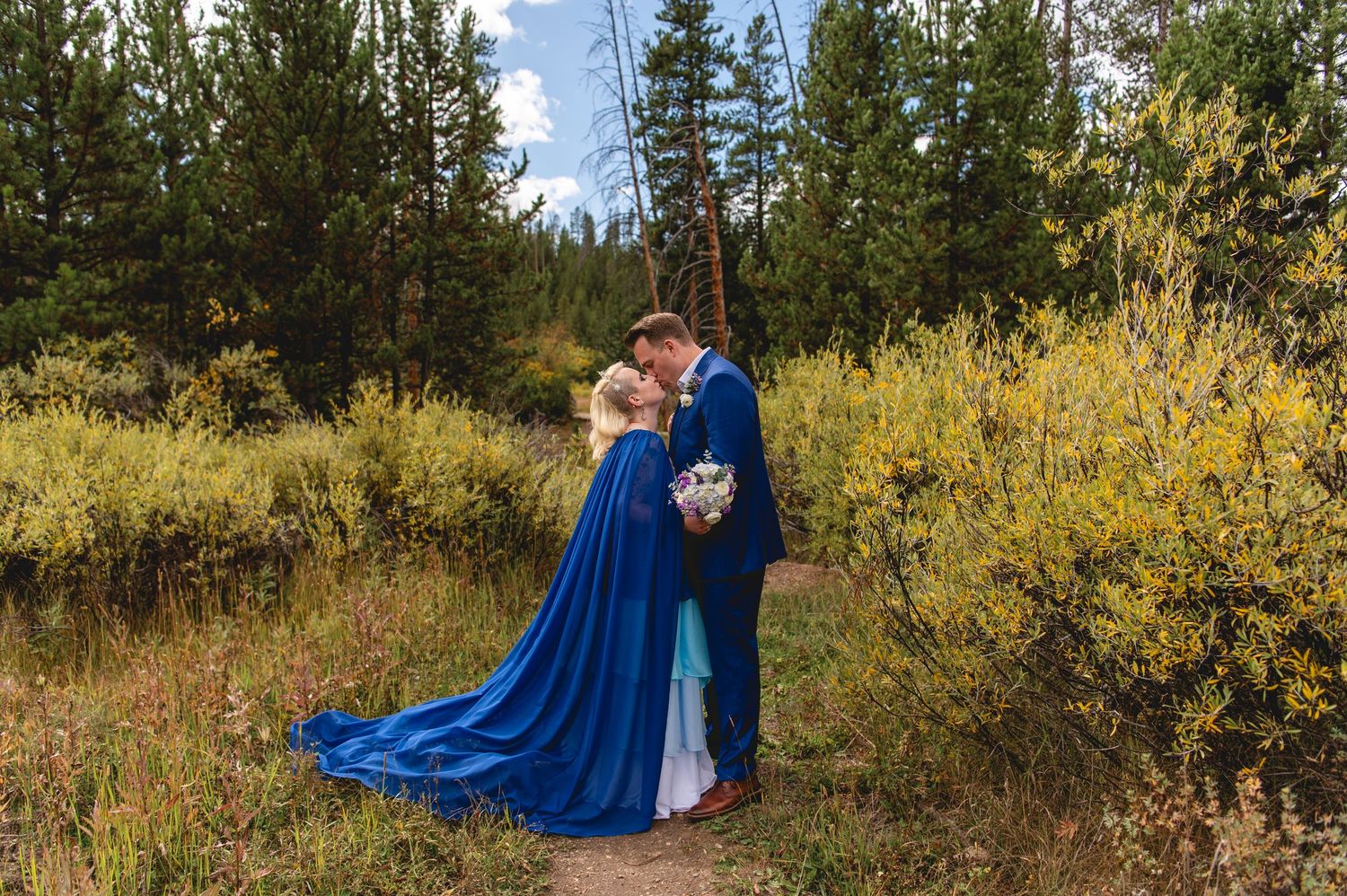 Couple in deep blue formal attire embraces against a backdrop of autumn foliage and evergreen trees in outdoor setting.