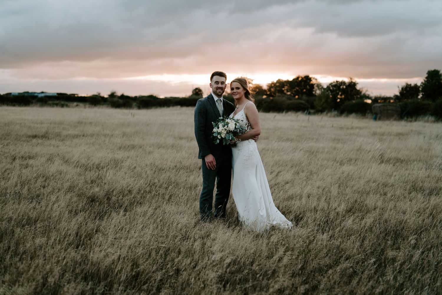 Wedding couple poses among lush greenery and white floral arrangements outdoors.