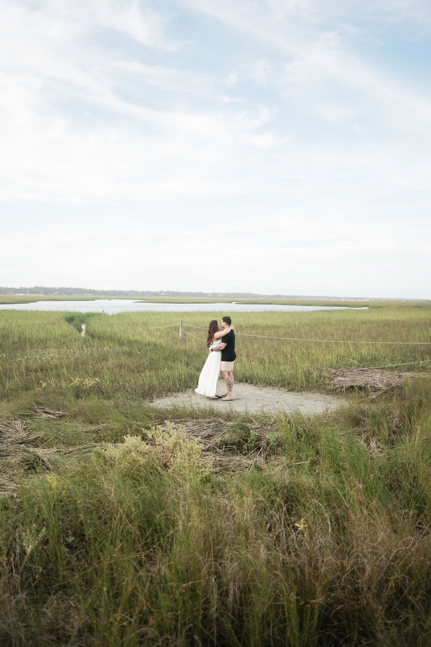A couple embraces on a small sandy patch surrounded by marsh grass and water on an overcast day.