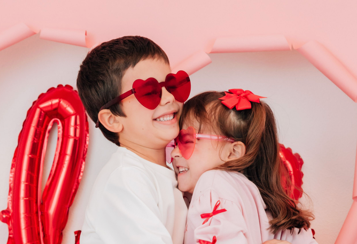 A boy and girl in heart-shaped sunglasses share a sweet cheek kiss with red love balloons nearby.