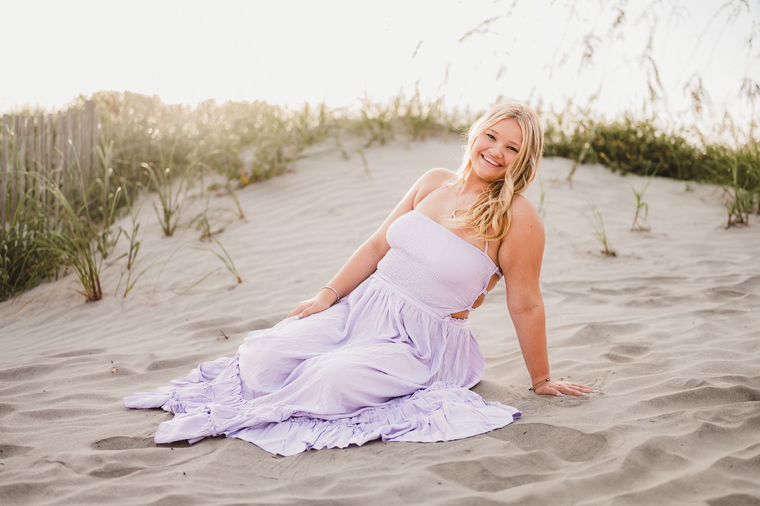 A person in a flowing lavender dress poses gracefully on sandy beach dunes during golden hour.