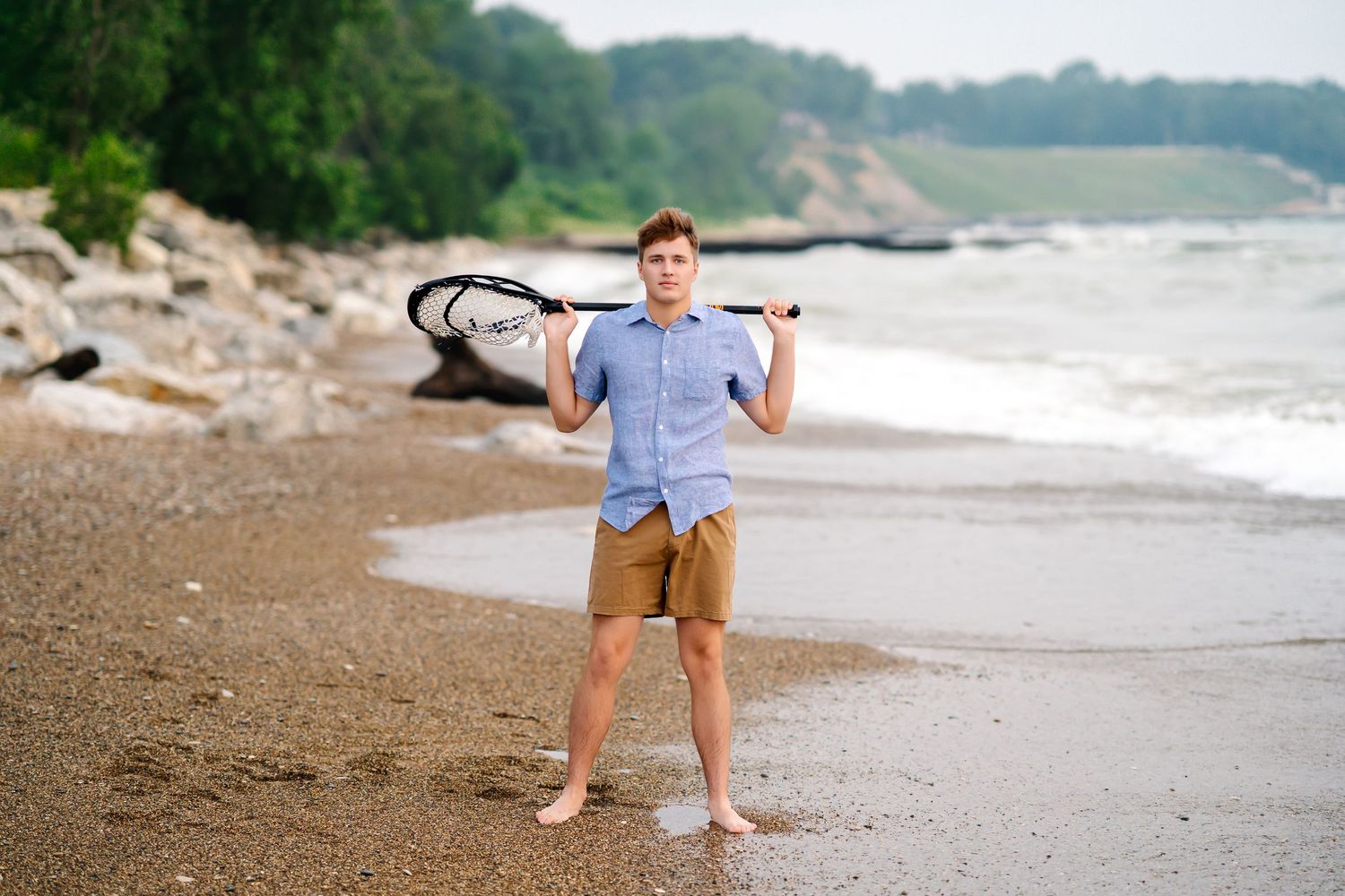 A person wearing business attire walks barefoot along a rocky beach shoreline on an overcast day.