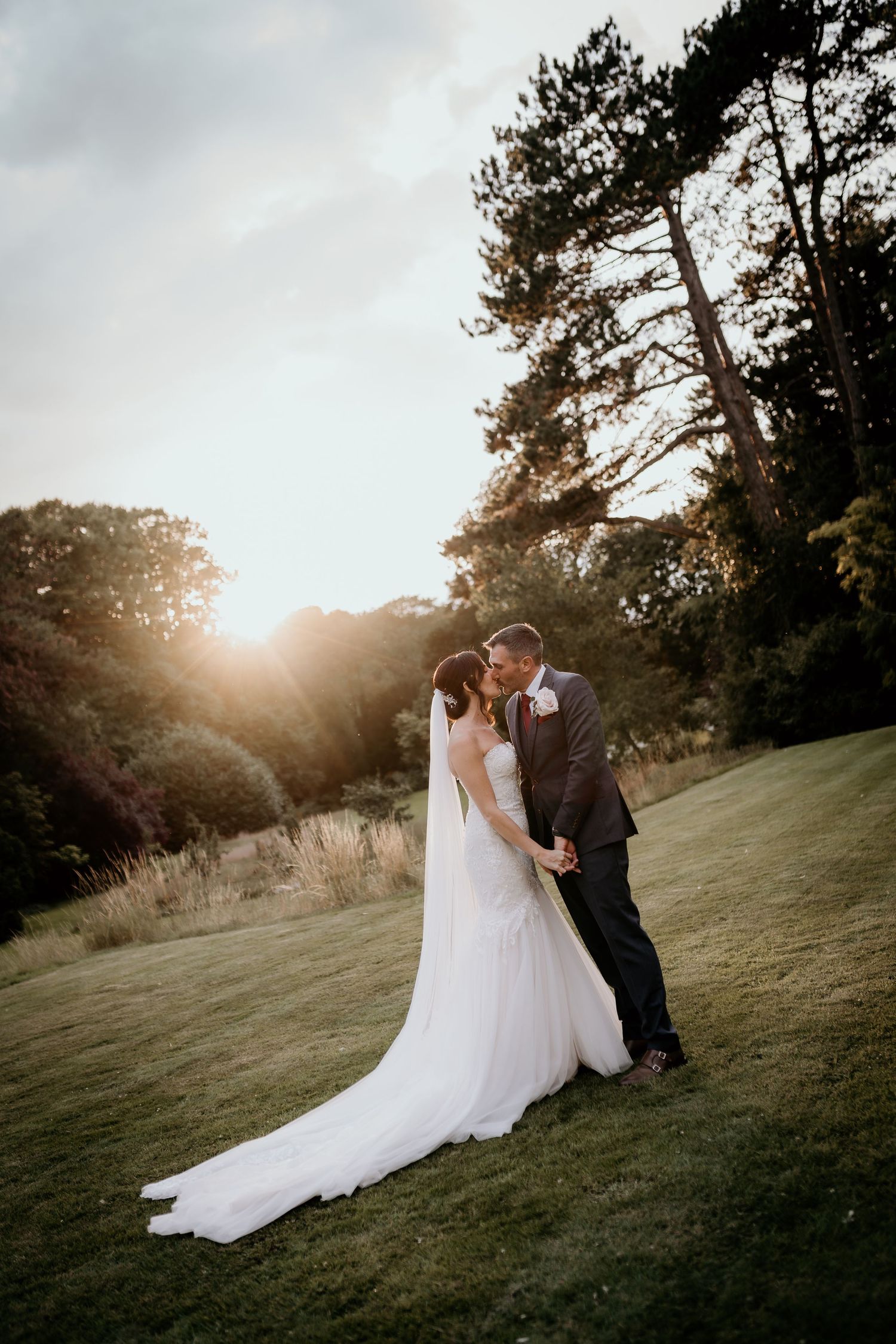 Newlywed couple sharing an intimate moment at sunset on a grassy hillside with tall trees in the background.