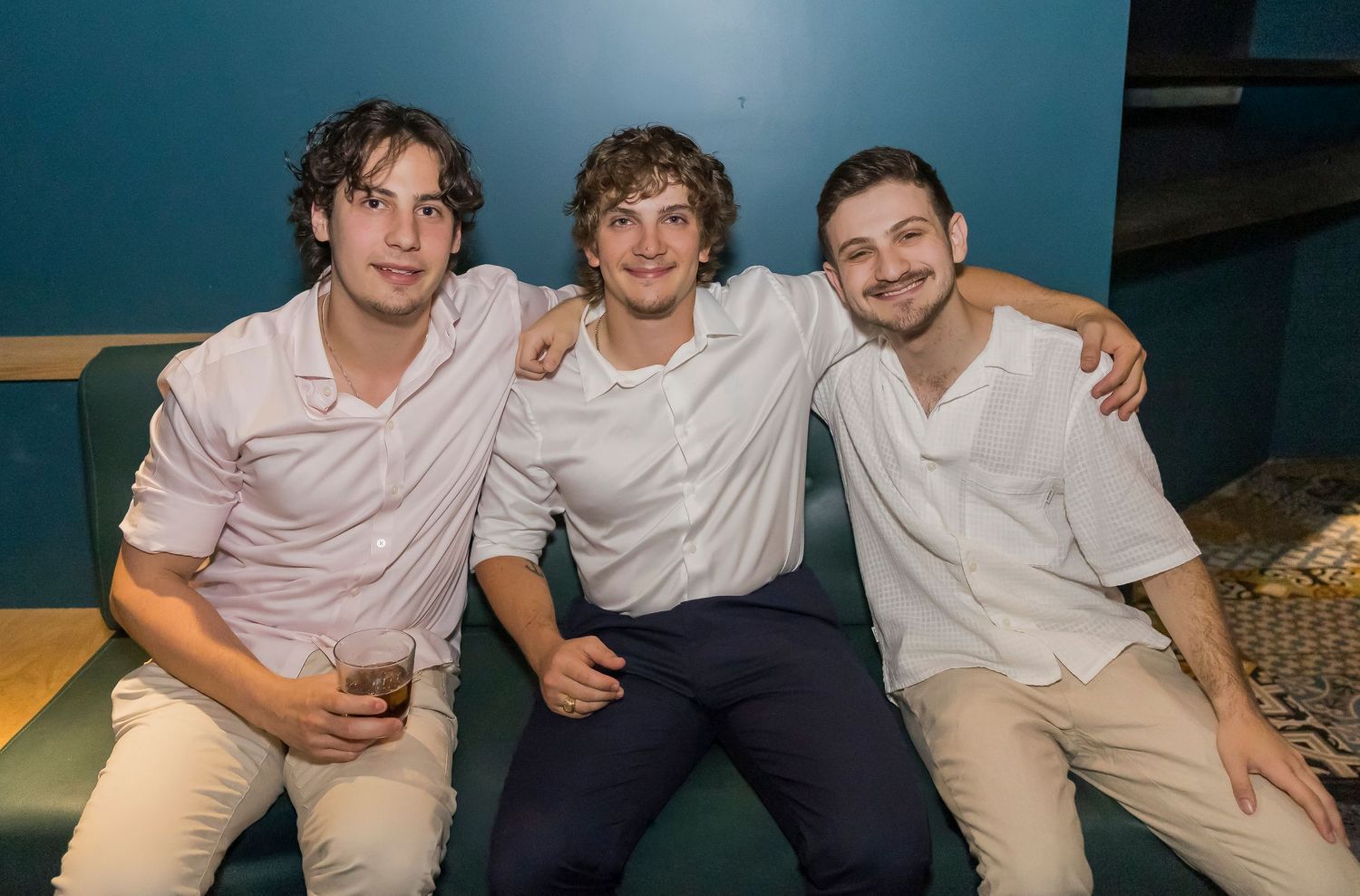 Three friends in casual white shirts and khaki pants sit together smiling on a couch.