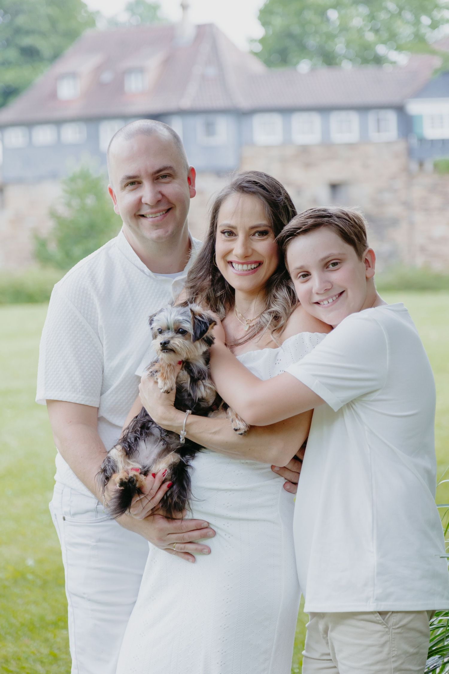 Family wearing white clothes relaxes together under shady trees in a summer garden setting.