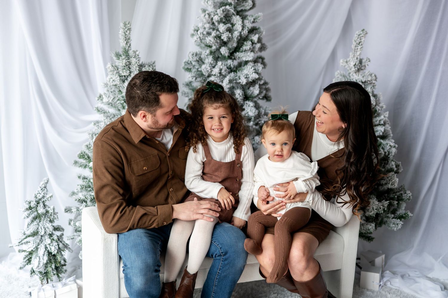 Family of four poses with snowy Christmas trees in white studio, parents gazing at two young daughters in brown and white outfits.