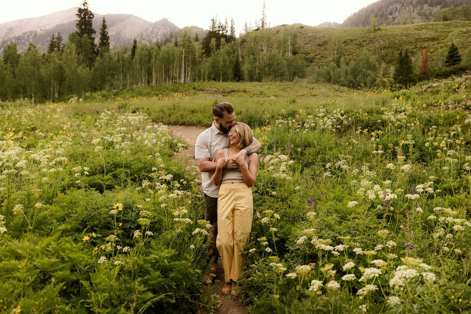 A couple embraces and laughs together in a golden field with mountains in the background during sunset.