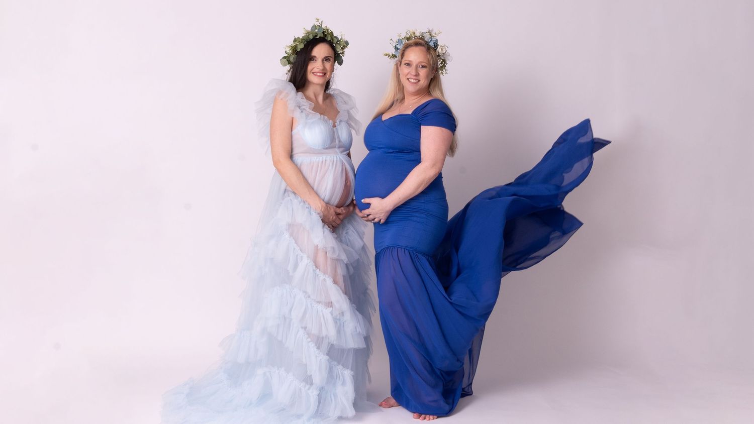 Two women in contrasting white and blue dresses pose together in a bright studio setting.