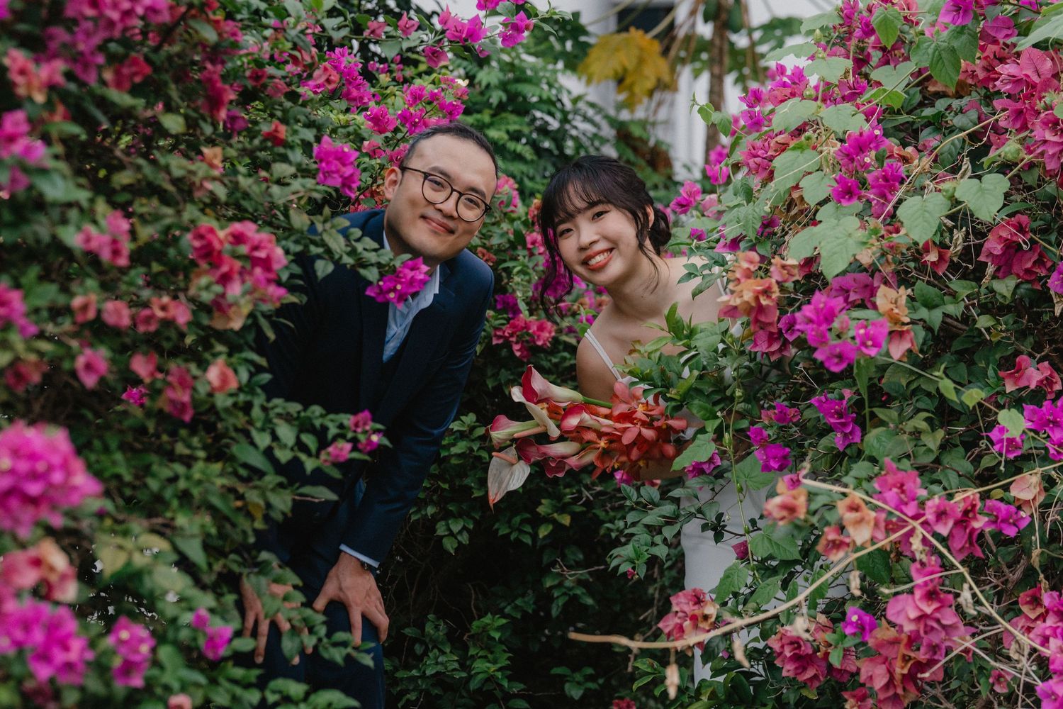A cheerful couple poses amongst vibrant pink bougainvillea flowers in a garden setting.