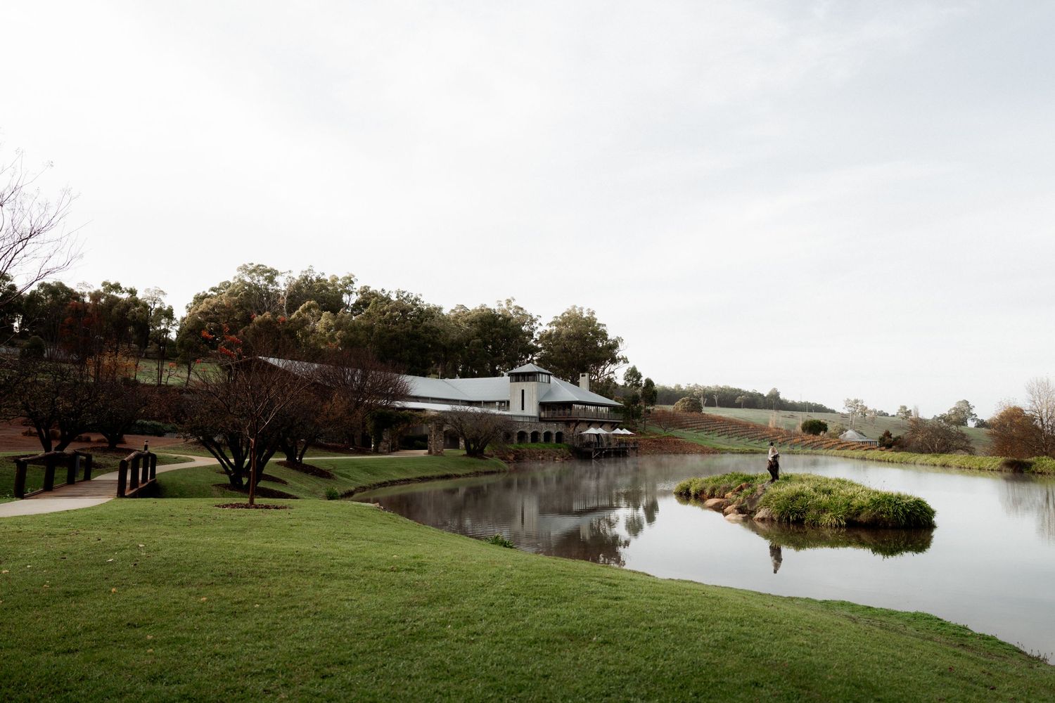 Modern building with expansive lawn sits beside a tranquil pond surrounded by trees on an overcast day.