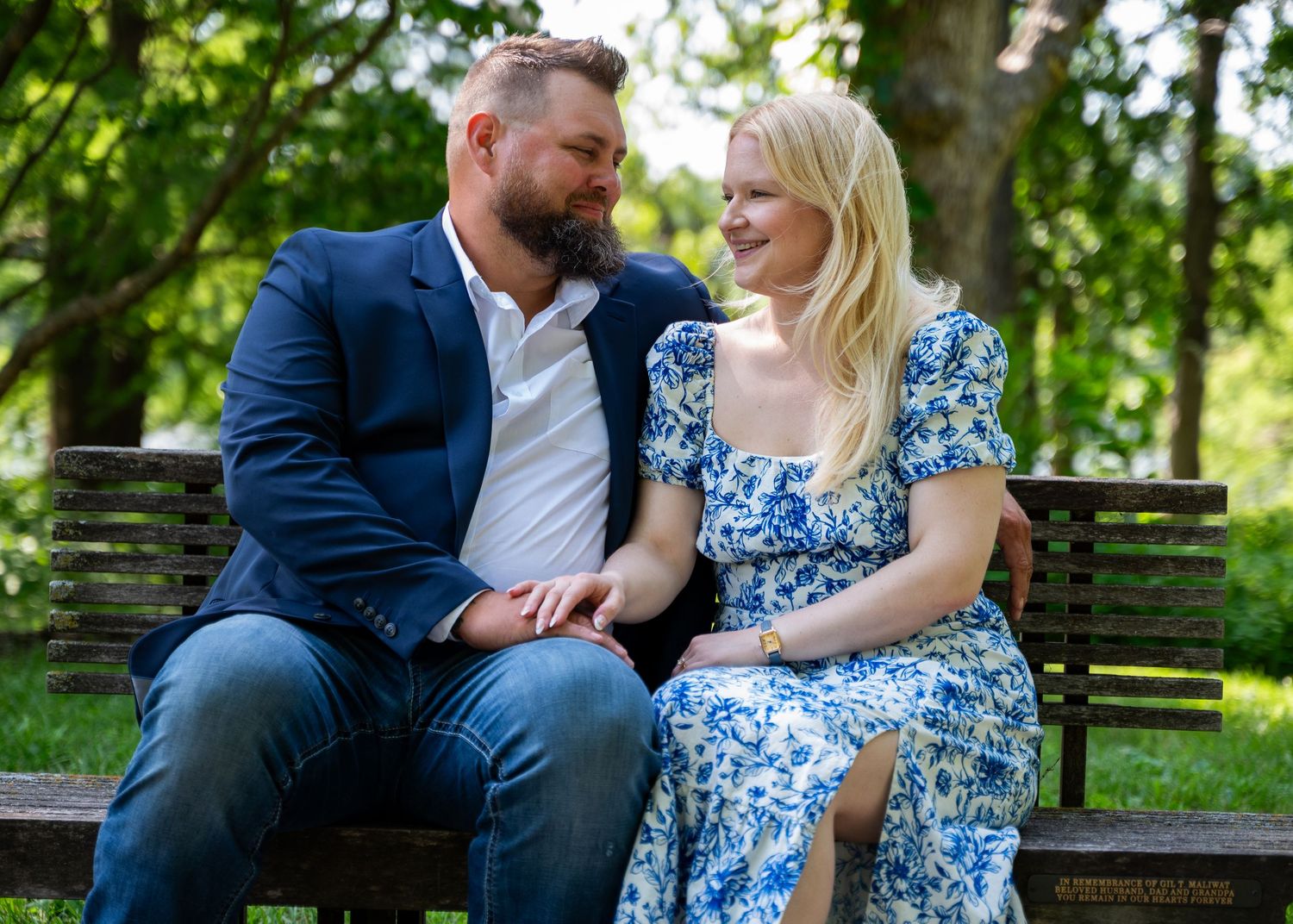 Engaged couple sitting together on bench in a garden lovingly looking at each other. Woman wearing flowy blue and white dress