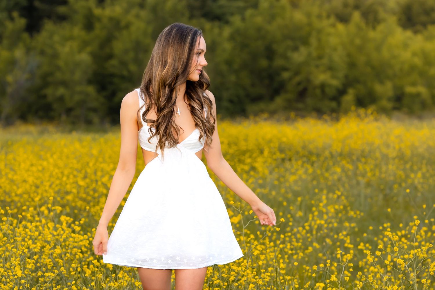 A young woman in a white sundress poses in a field of bright yellow wildflowers on a sunny summer day.