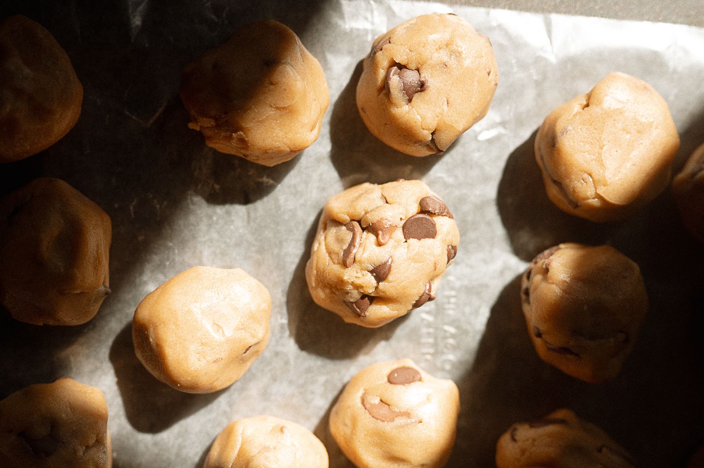 Raw chocolate chip cookie dough balls arranged on parchment paper with dramatic lighting.
