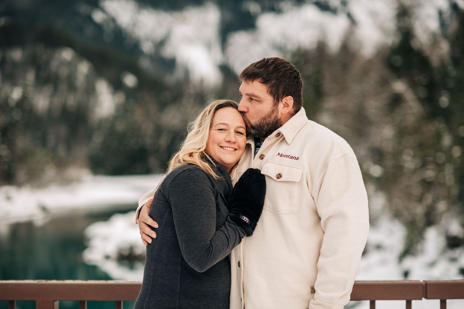 A couple shares an intimate moment on a deck overlooking a snowy winter landscape with a lake in the background.