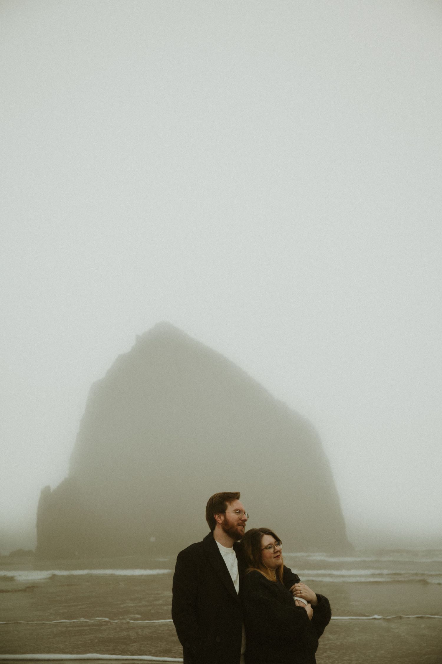 Two figures stand silhouetted against dramatic rocky cliffs during golden hour creating a moody coastal landscape scene.