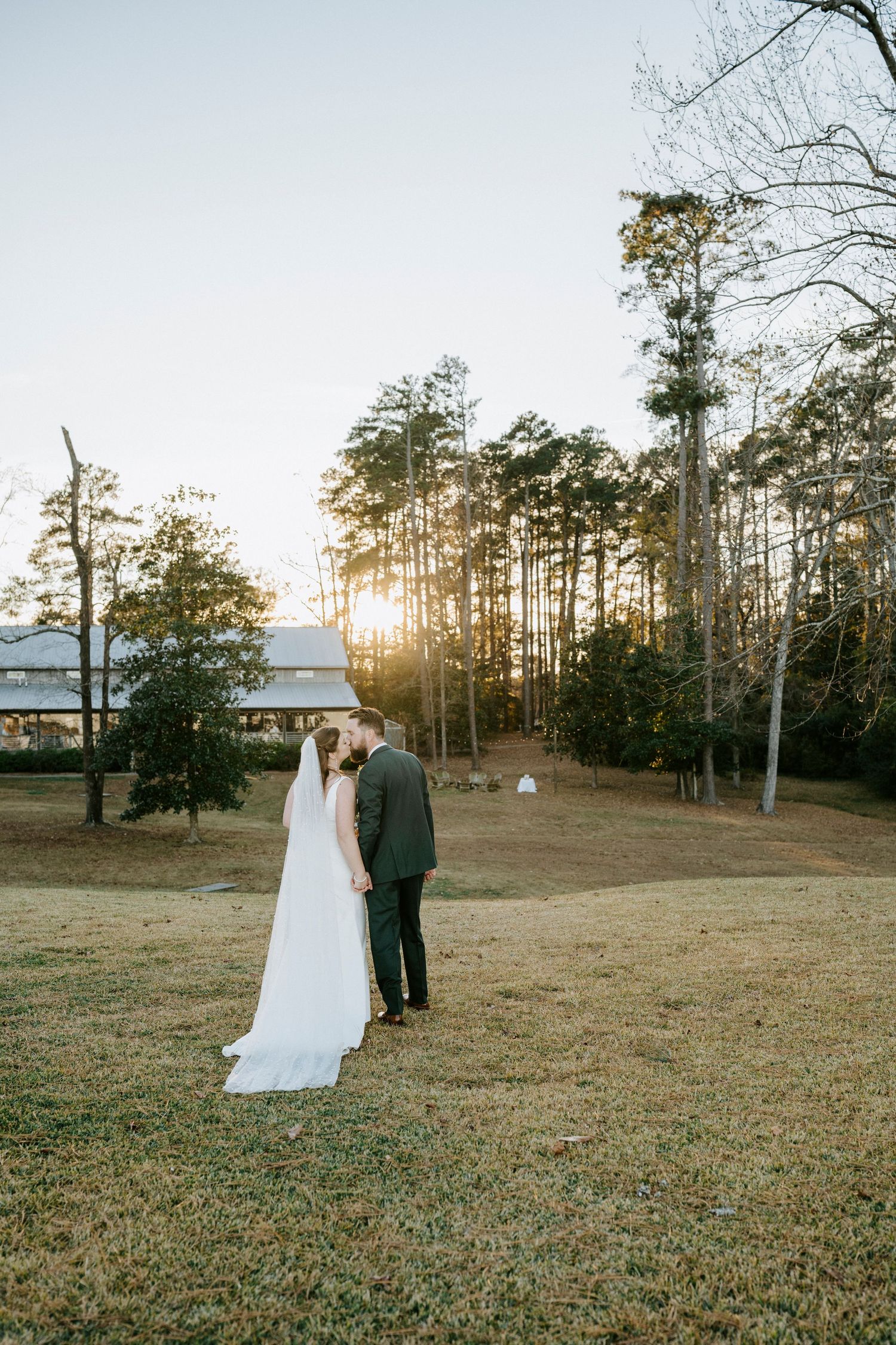 A couple in wedding attire stands together in a rustic outdoor field at sunset with pine trees in the background.