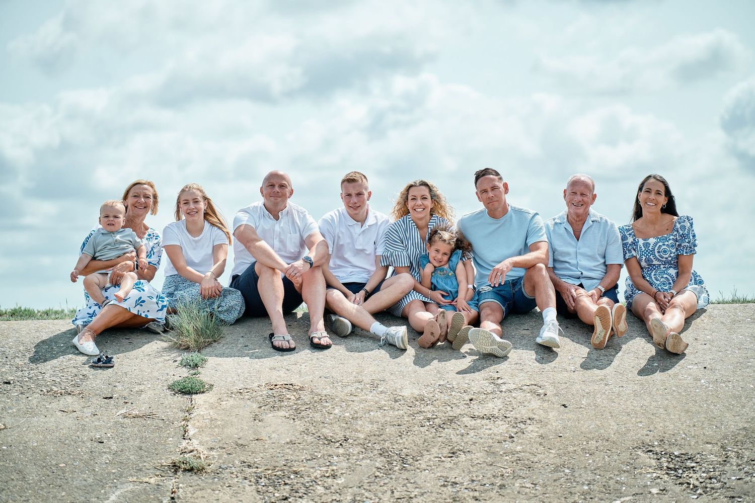 A multigenerational family group wearing coordinating blue outfits sits together on a beach on a cloudy day.