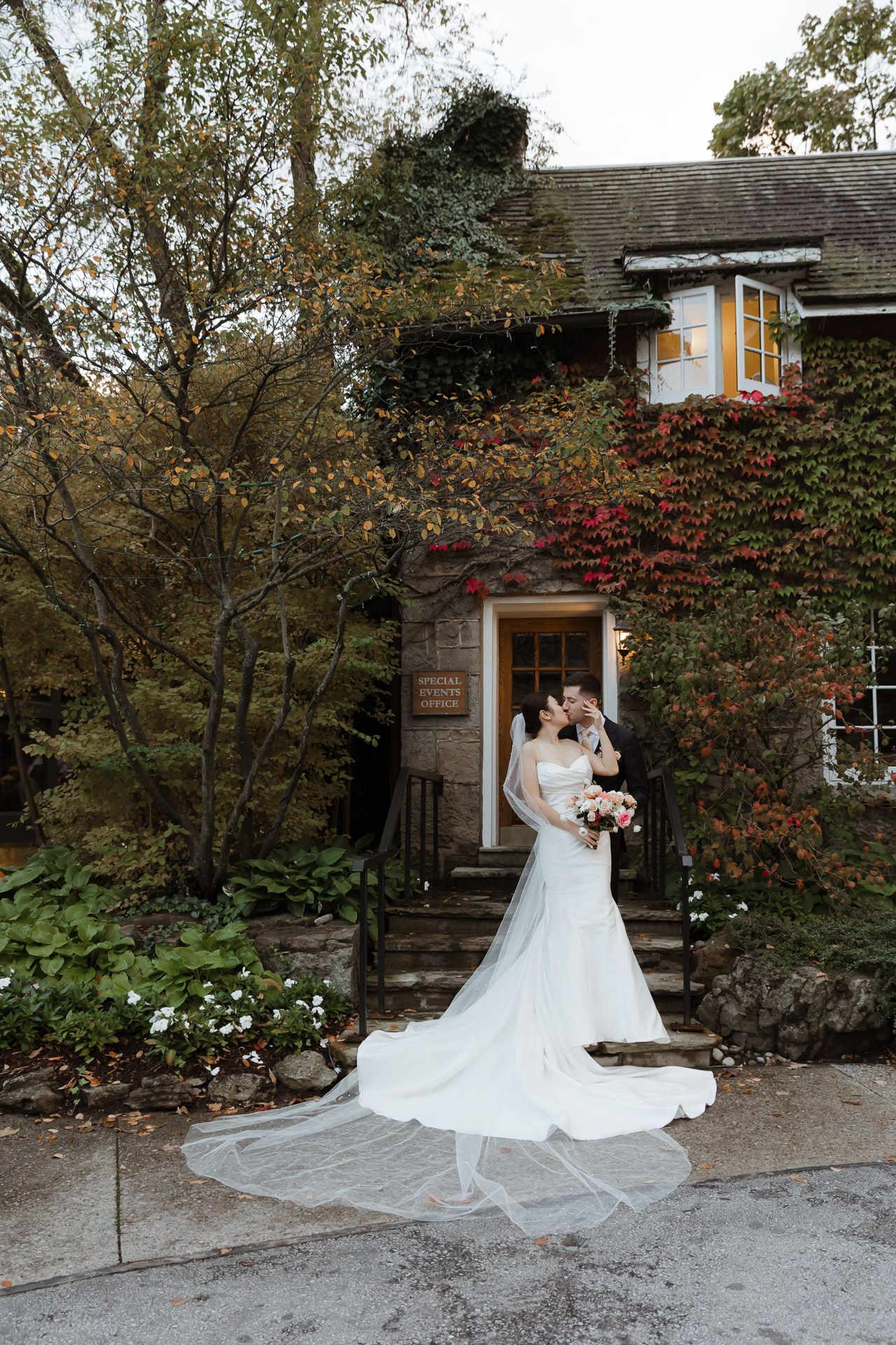 A couple in formal wedding attire holds hands while standing on a city street corner with historic brick buildings in the background.