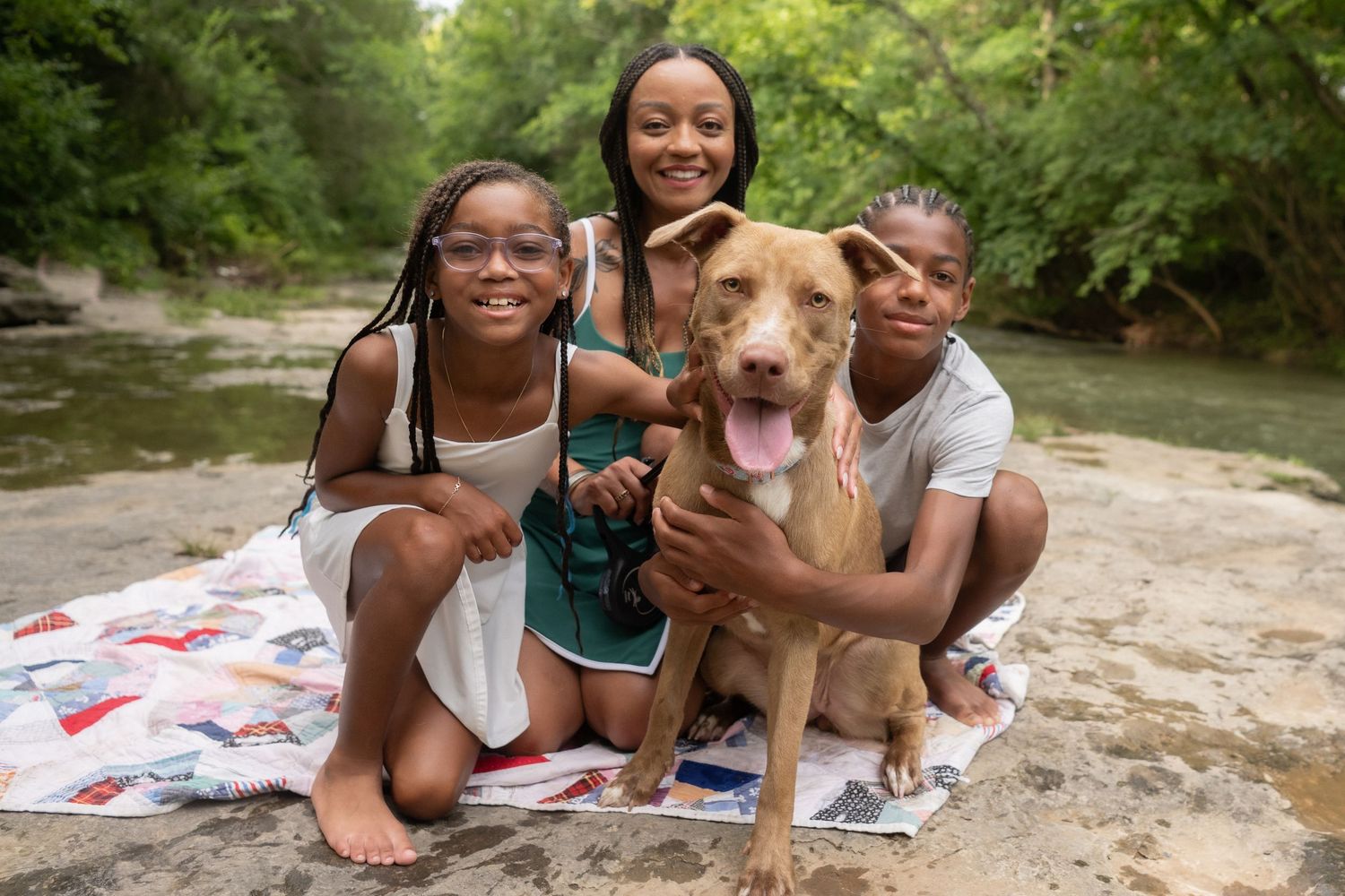 A group takes selfie photos with a happy tan pit bull type dog outdoors on a sunny day.