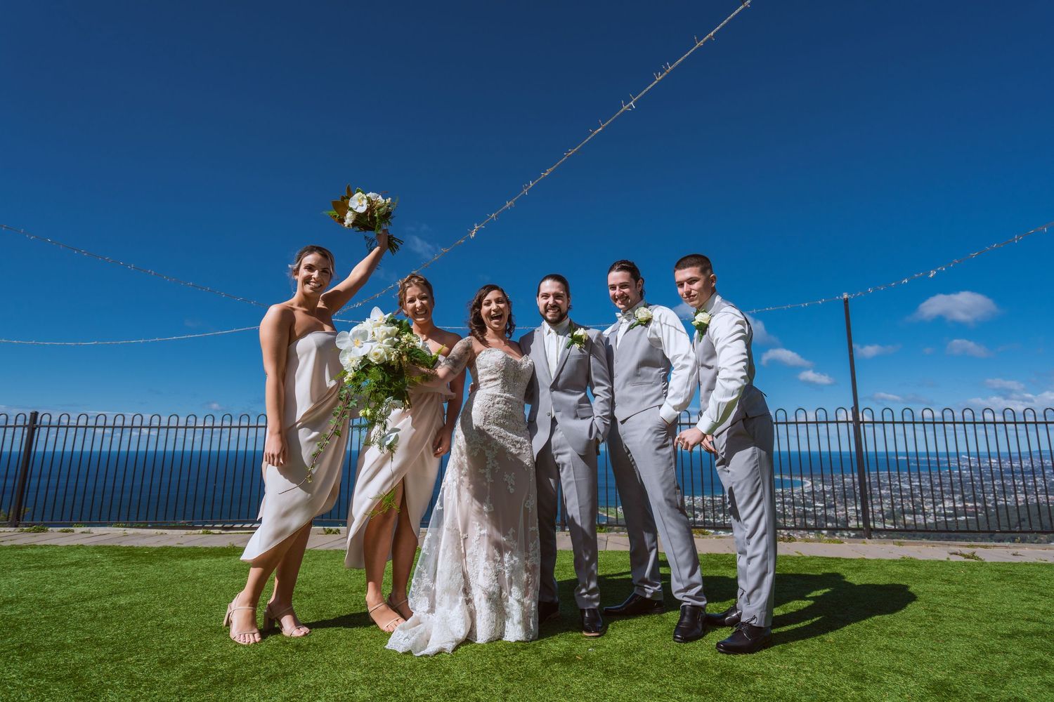 Wedding party in white attire poses playfully on grassy hilltop against bright blue sky with ocean view.