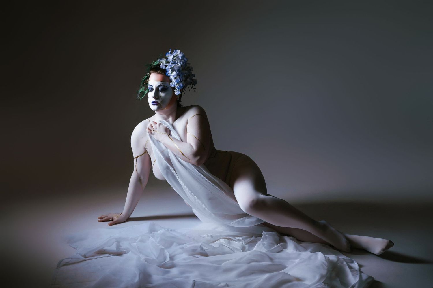 A graceful figure in white fabric reclines on a dark floor creating an ethereal artistic composition.