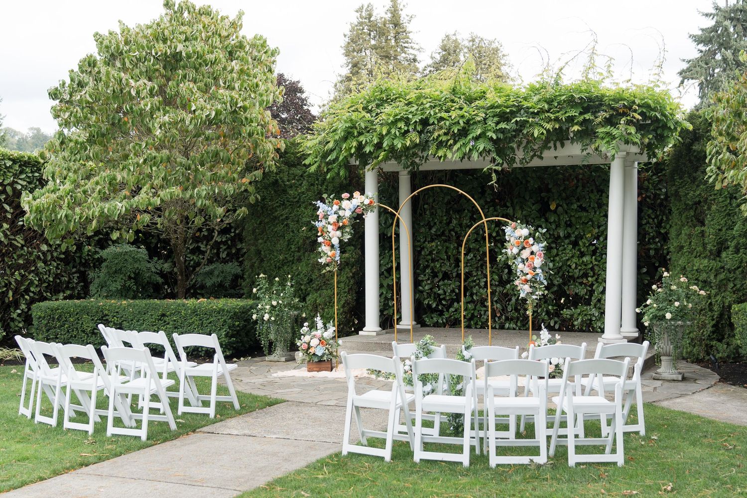 Elegant outdoor wedding ceremony setup with white chairs, floral arch, and ivy-covered pergola in a lush garden.