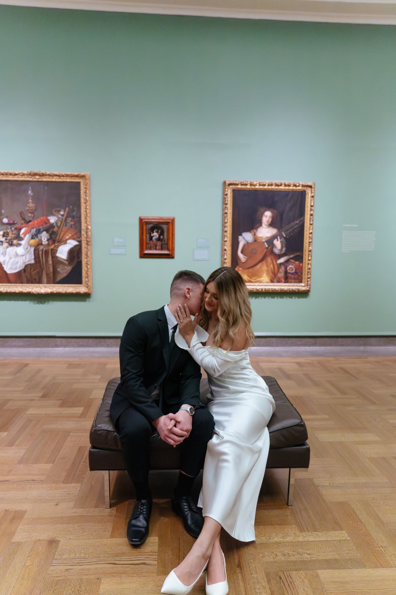 A romantic couple shares an intimate moment on a museum bench with classical paintings displayed on mint green walls.