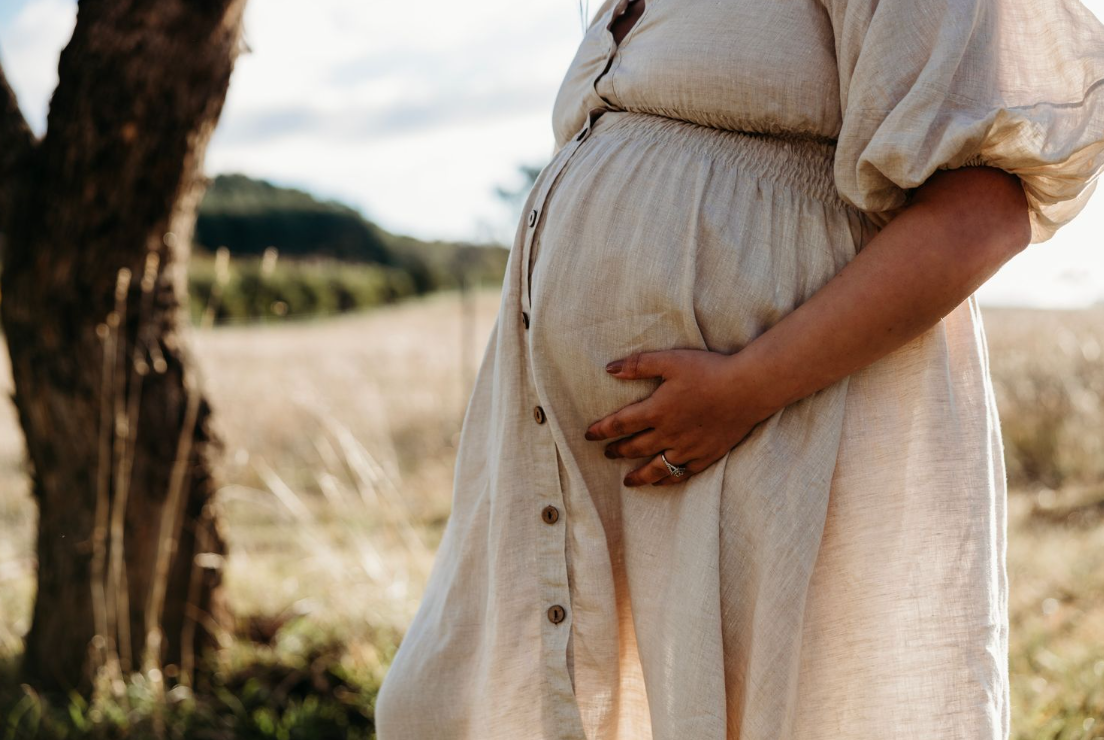 Close-up of pregnant belly in flowing white dress against rural landscape.