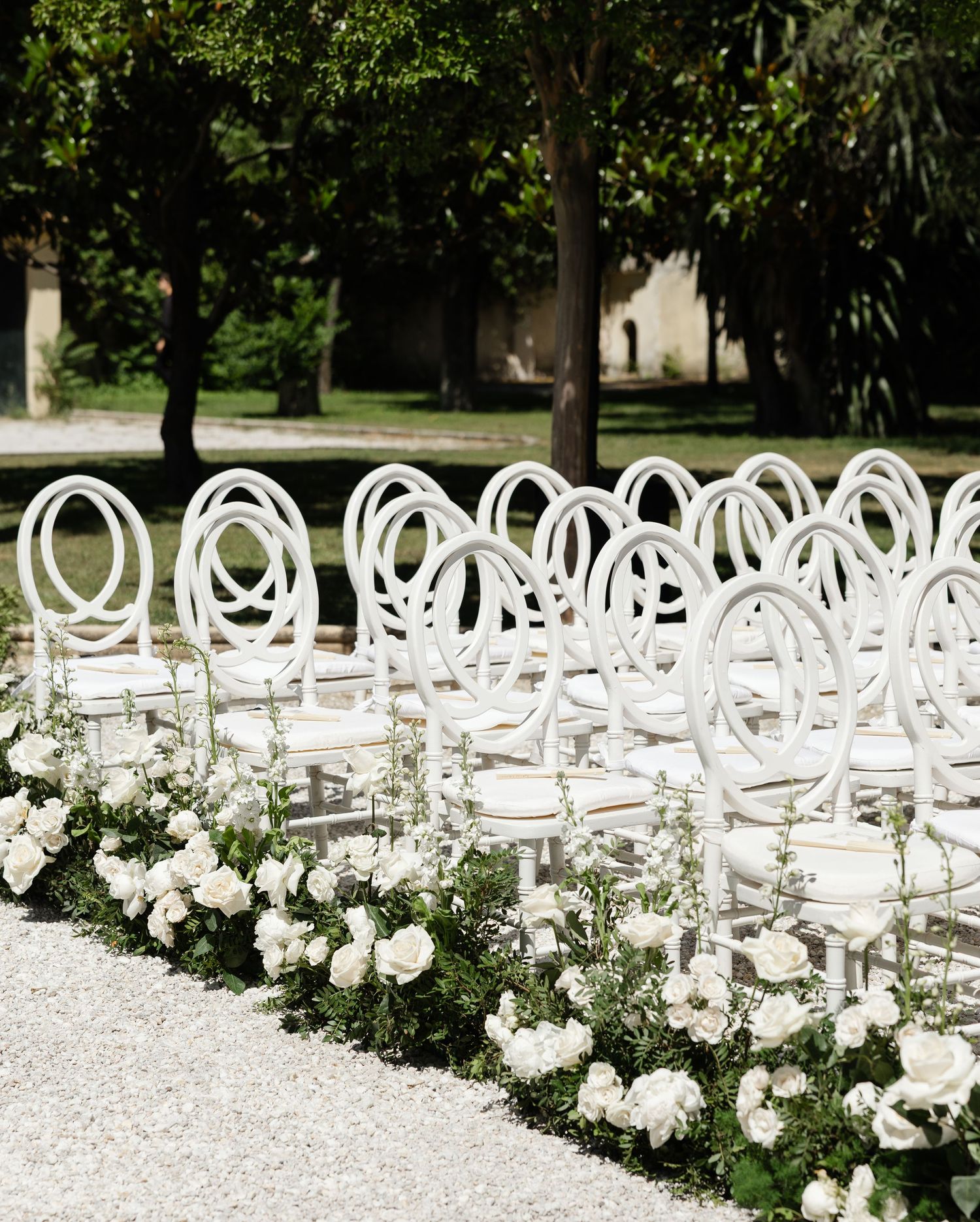 White circular wedding ceremony chairs arranged in rows with white floral arrangements lining the aisle in an outdoor garden setting.