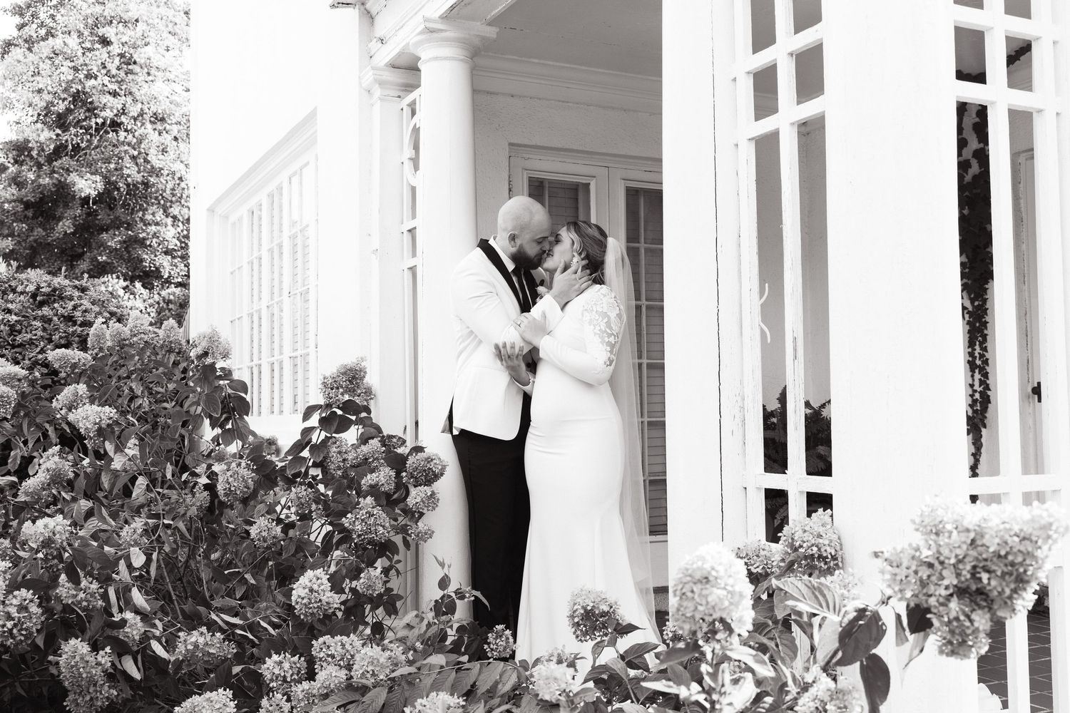 Black and white photo of an outdoor garden wedding ceremony with guests seated in white chairs facing a floral arch.