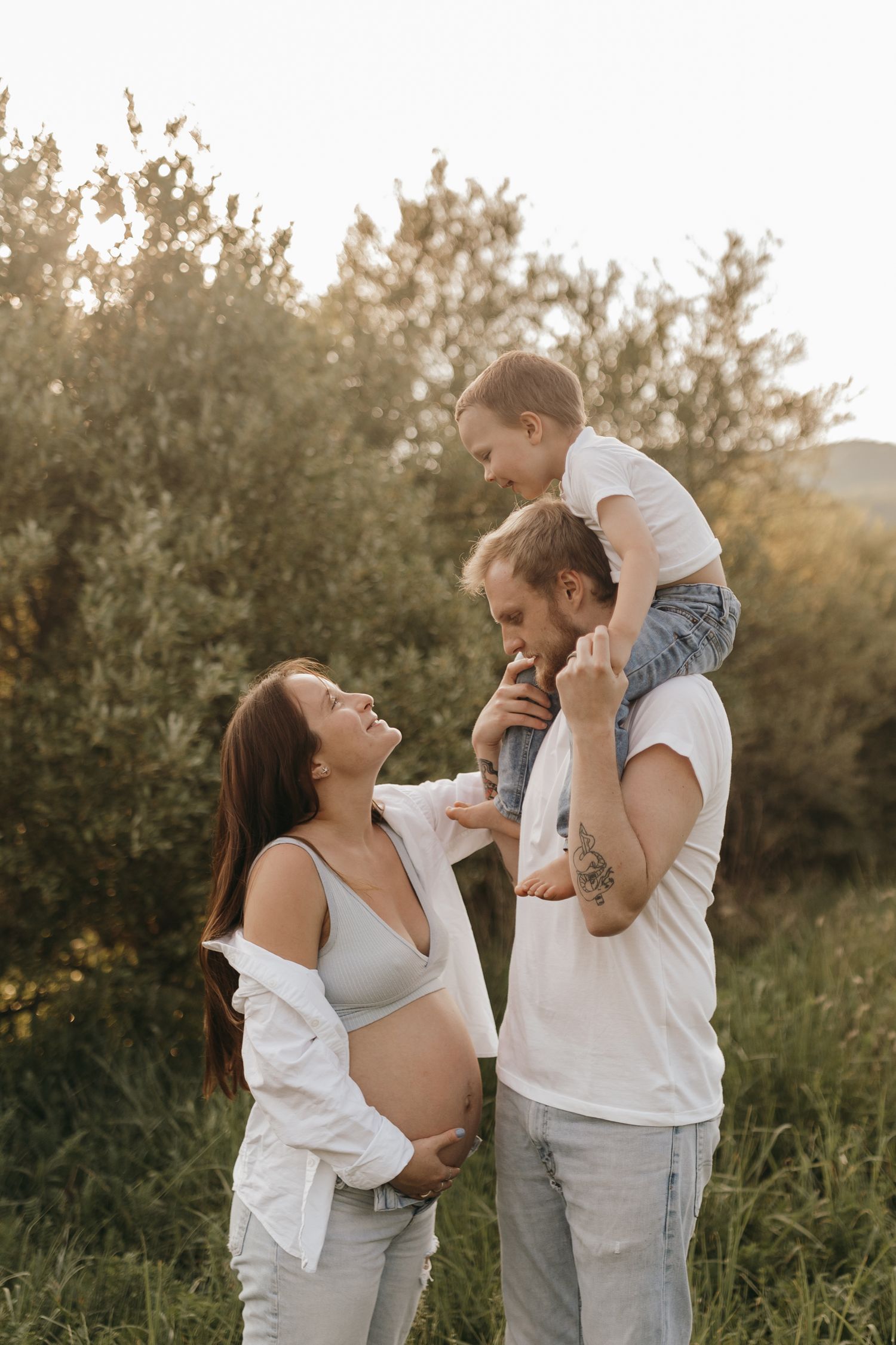 A joyful outdoor family moment at sunset with a pregnant woman and child on shoulders.